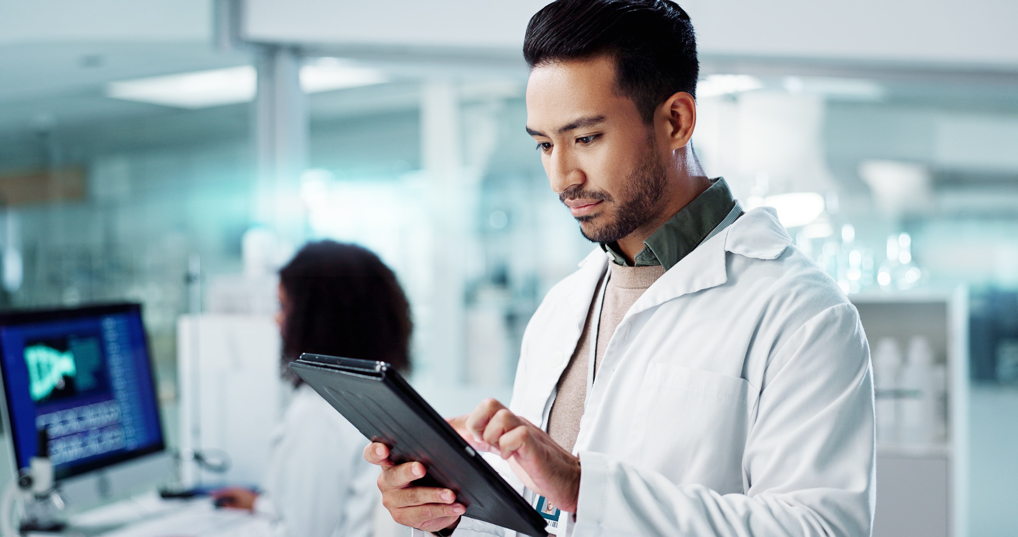 A lab technician in a white coat engages with a tablet while working in a laboratory.