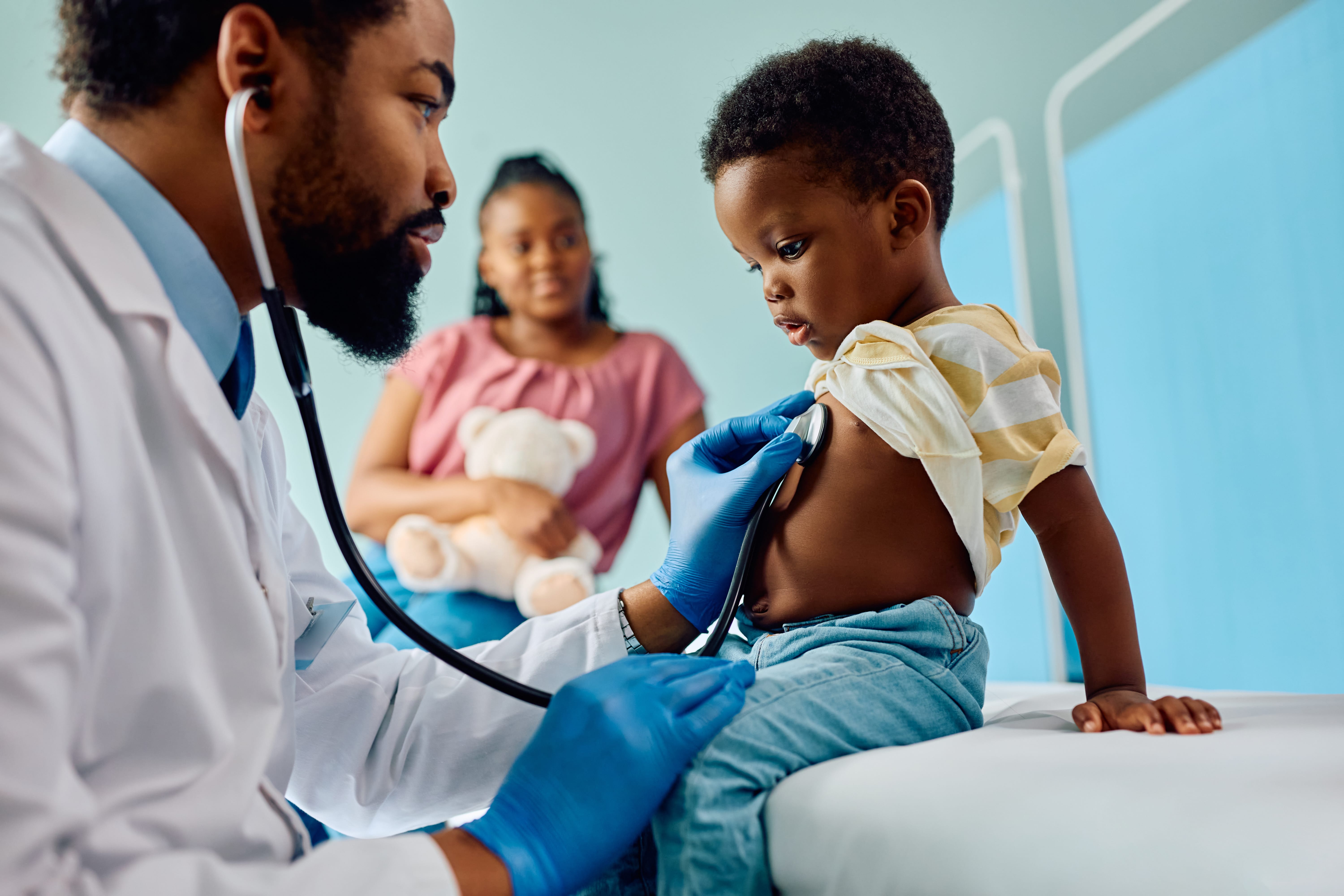 A doctor using a stethoscope to examine a young child in a medical office, with a parent present.
