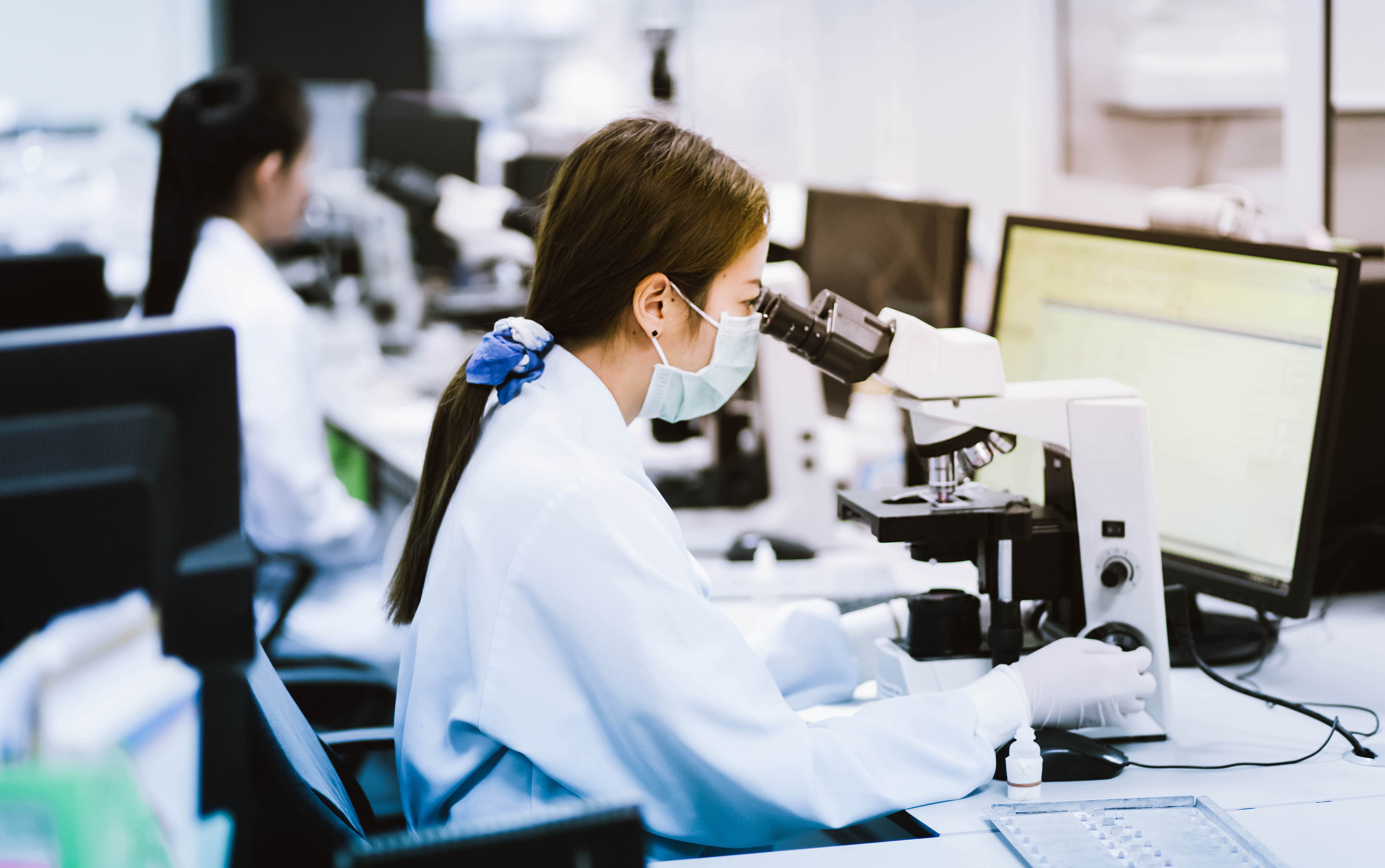 Female researcher wearing a mask and gloves examining samples under a microscope in a modern lab.