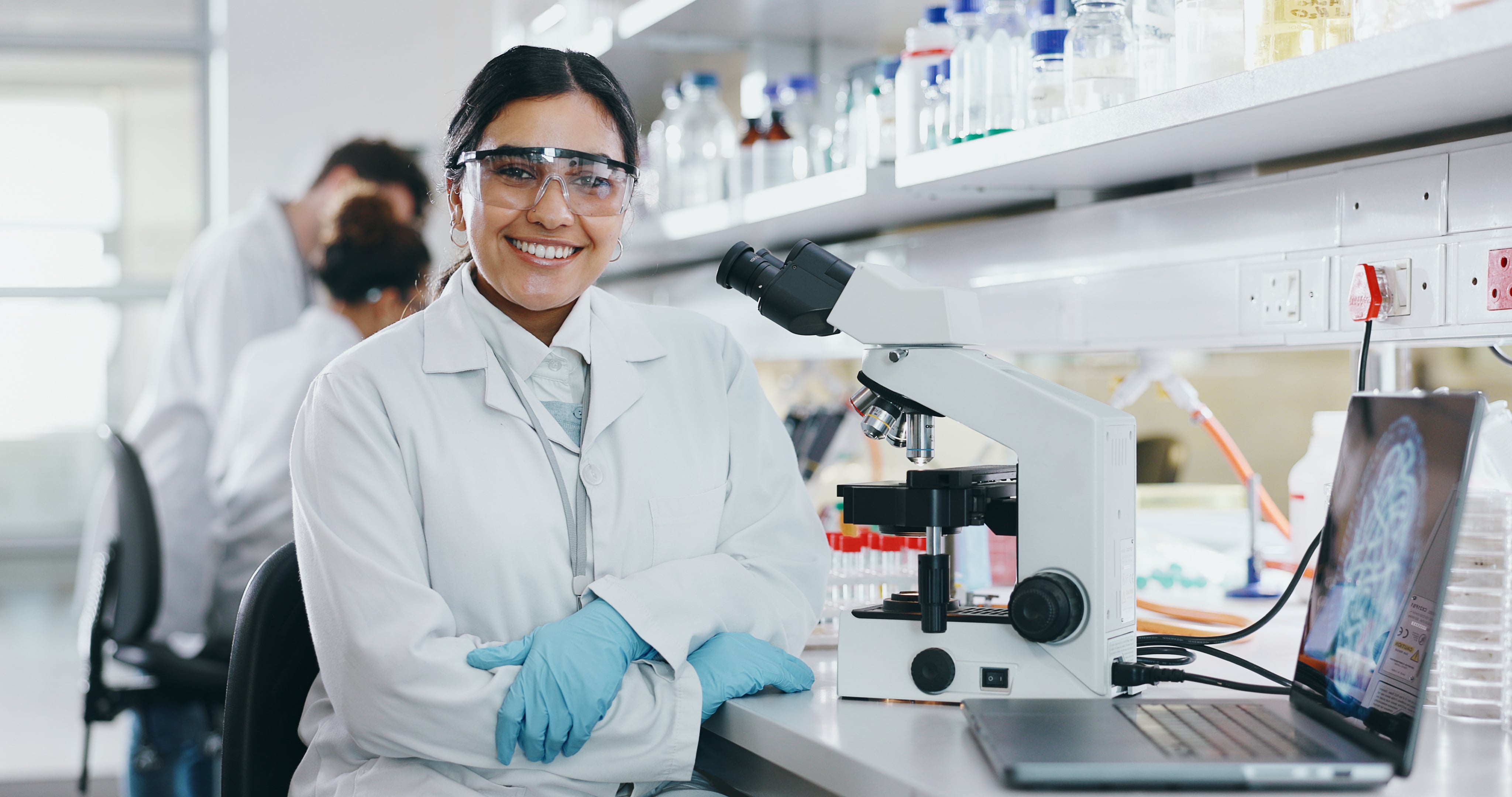 Female scientist wearing a lab coat and goggles, standing next to a microscope in a laboratory setting.