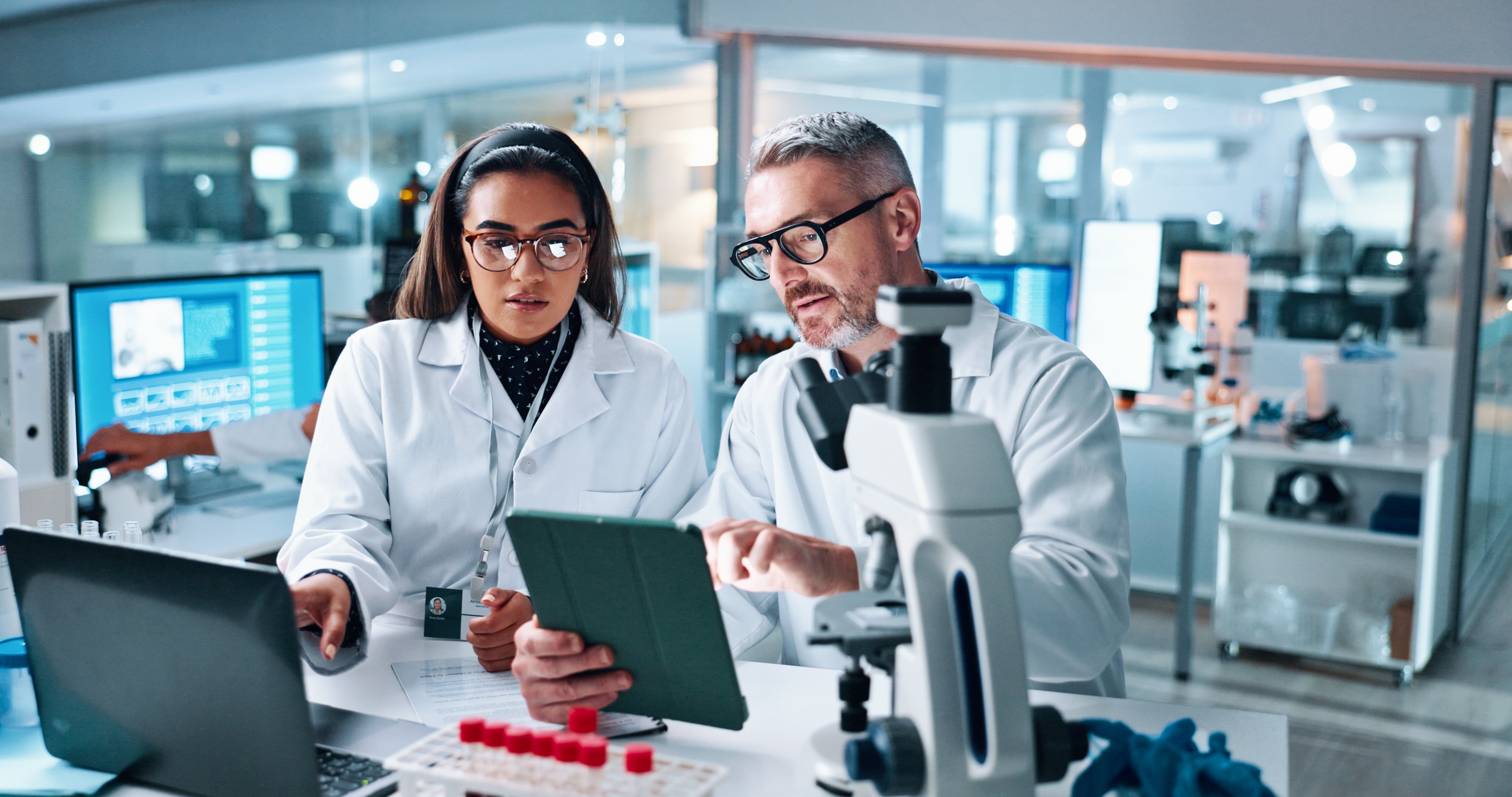Two scientists discussing research findings on a tablet in a lab environment, with laboratory equipment in the background.