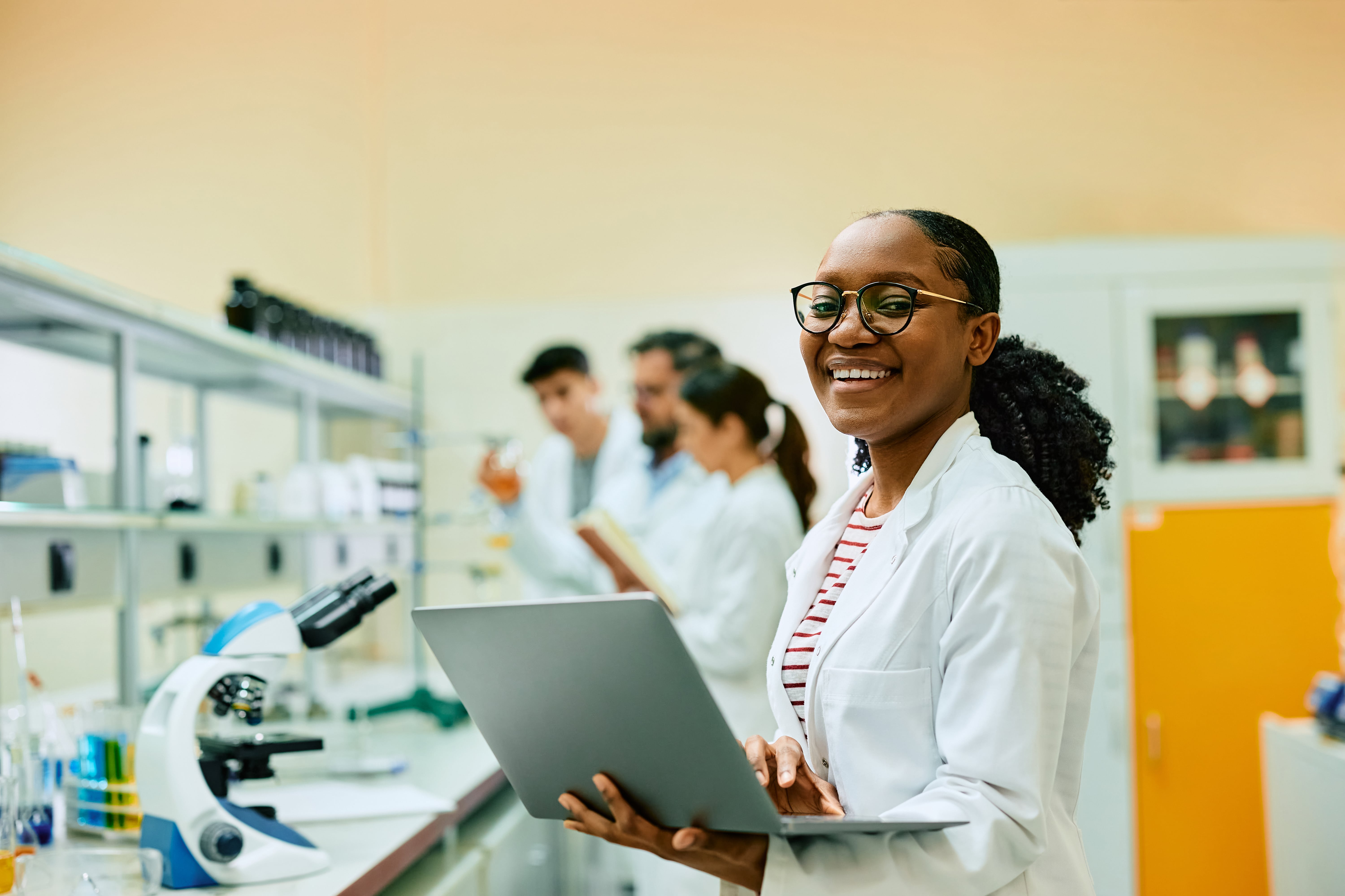 Smiling scientist in a lab coat holding a laptop, with colleagues in the background conducting experiments.