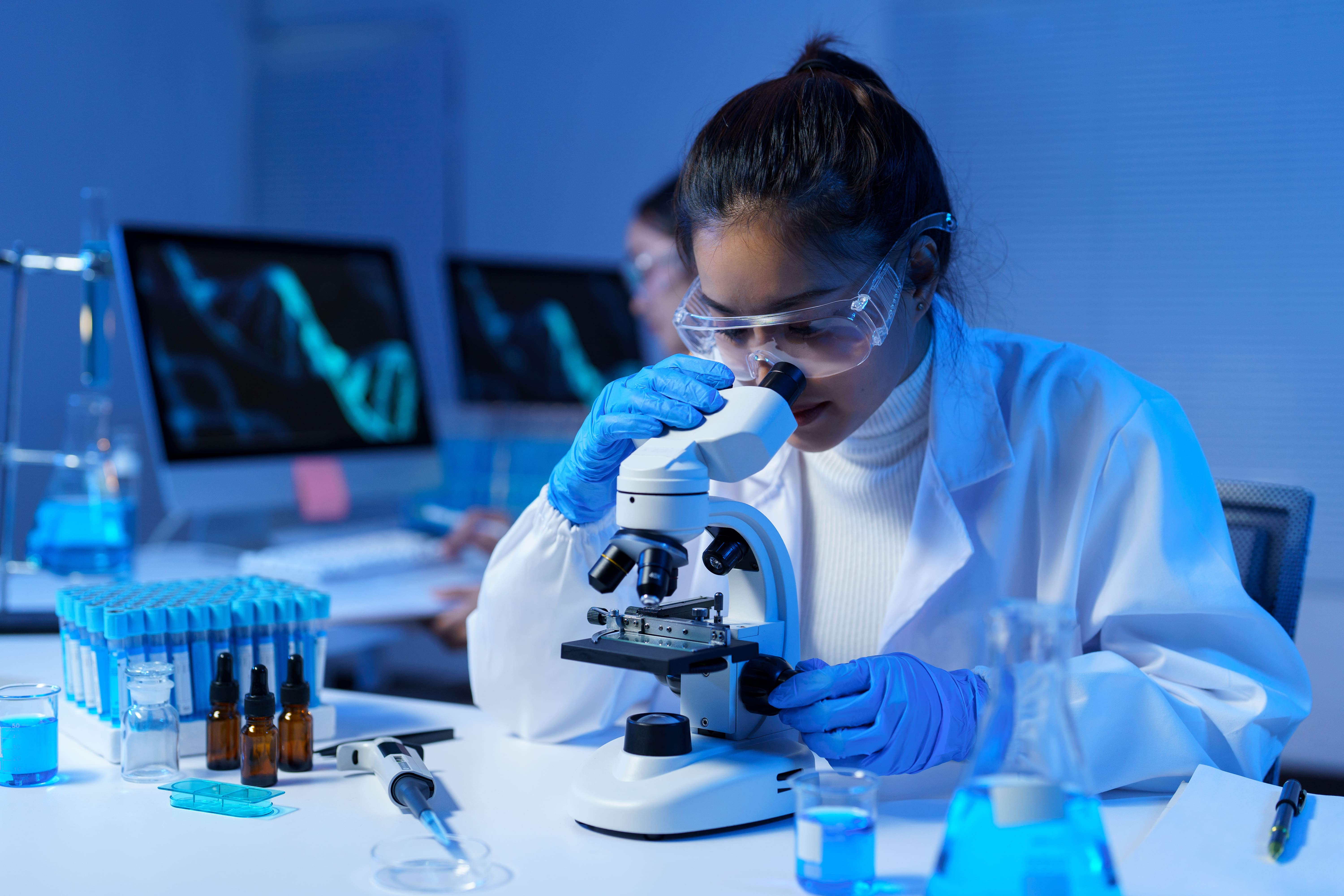 Female researcher wearing a lab coat and goggles using a microscope, with blue chemical solutions and equipment in the background