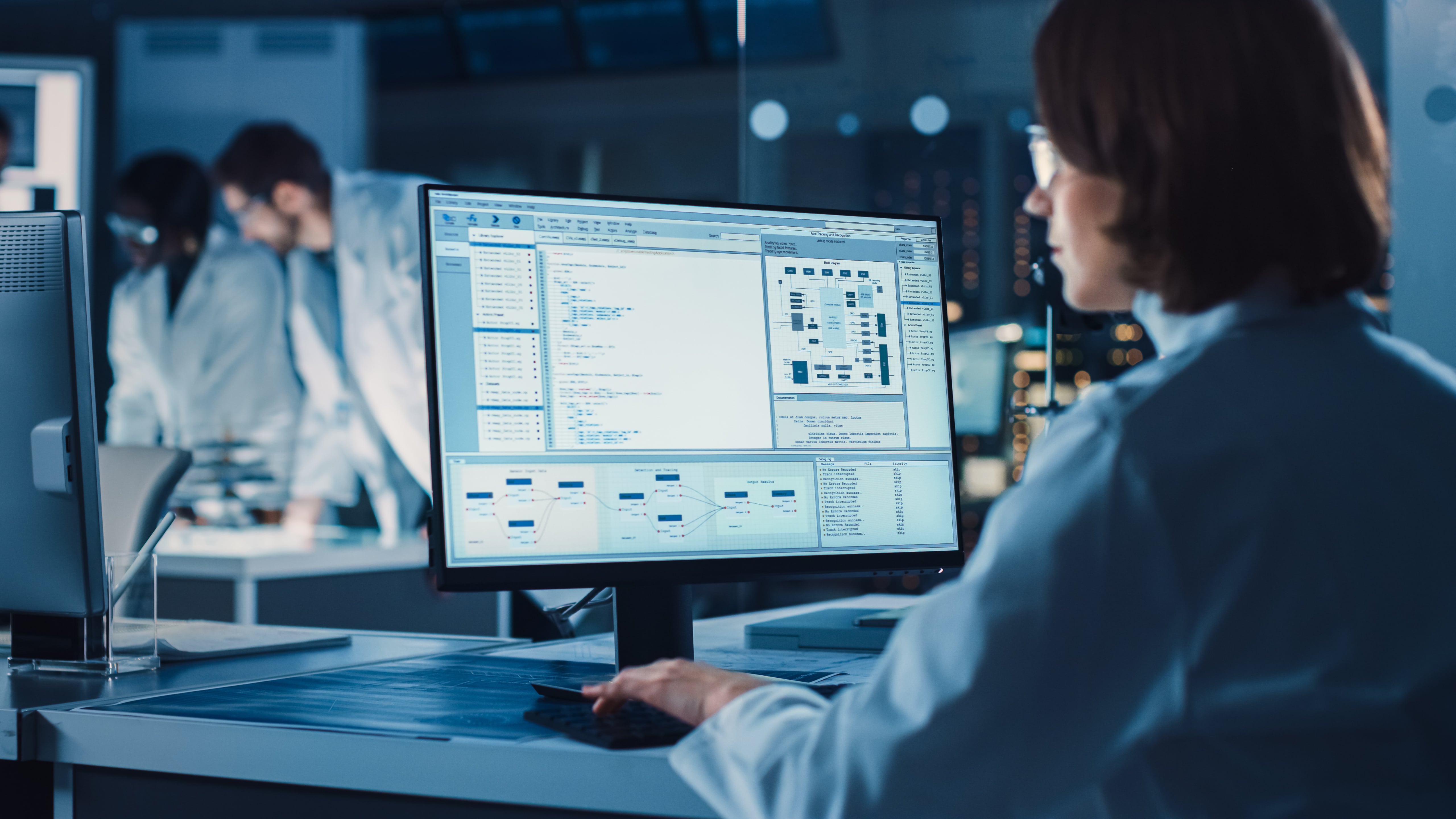 A female scientist focused on a computer screen displaying data and analysis, with colleagues in the background in a laboratory setting.