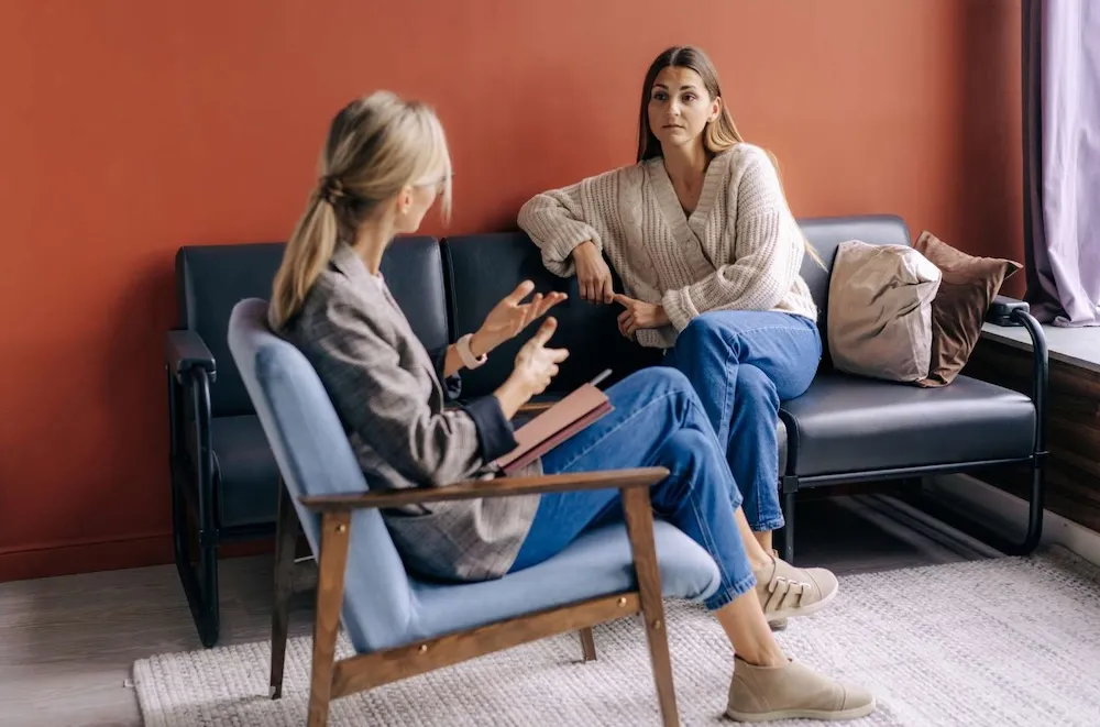 A speech-language pathologist speaks with a woman during a one-on-one session in a calm, comfortable room. The client listens attentively while seated on a couch, suggesting a thoughtful exchange.