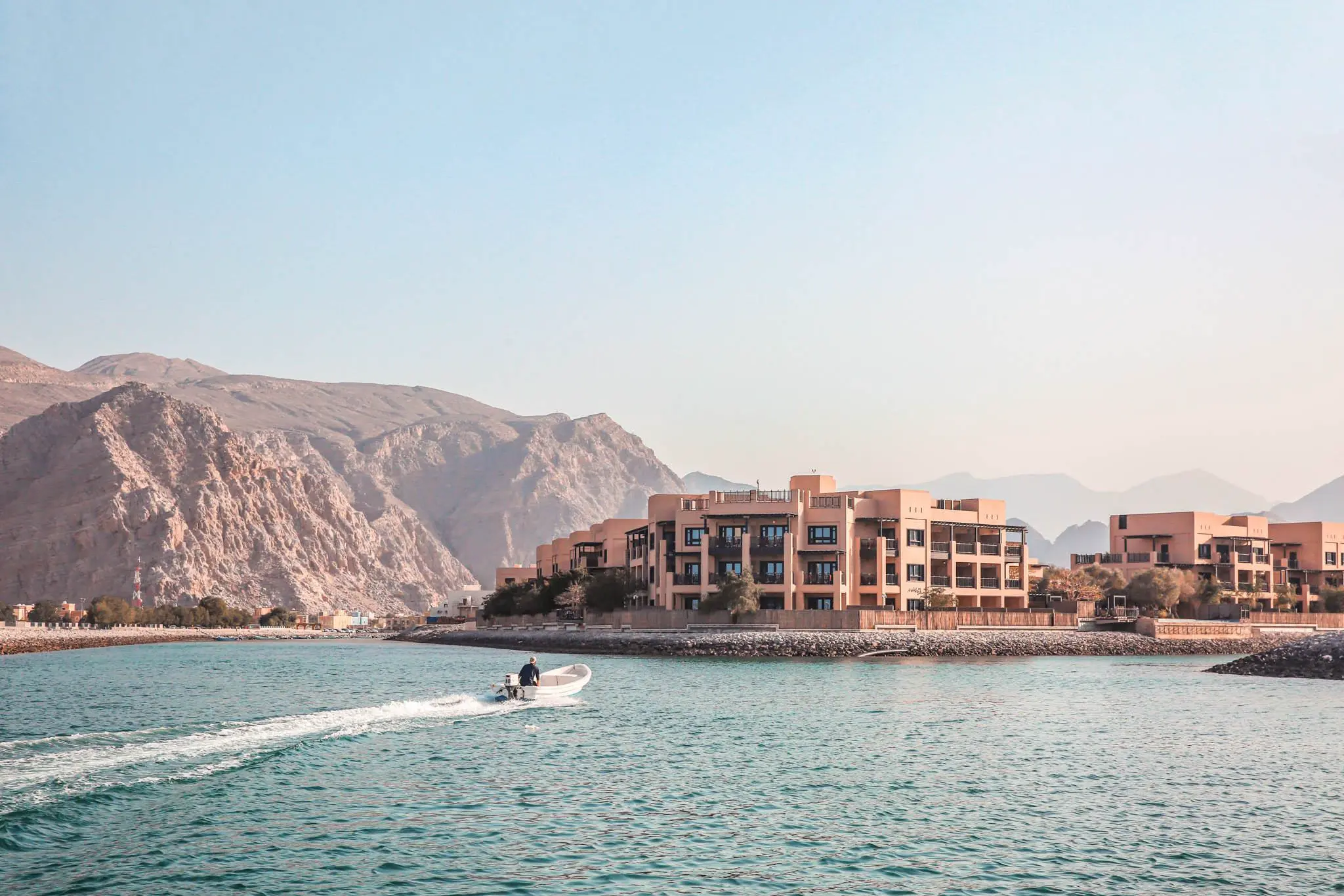 Atana Musandam property image taken from the water with the mountains in the background. A small speedboat rides in from the left of the image.