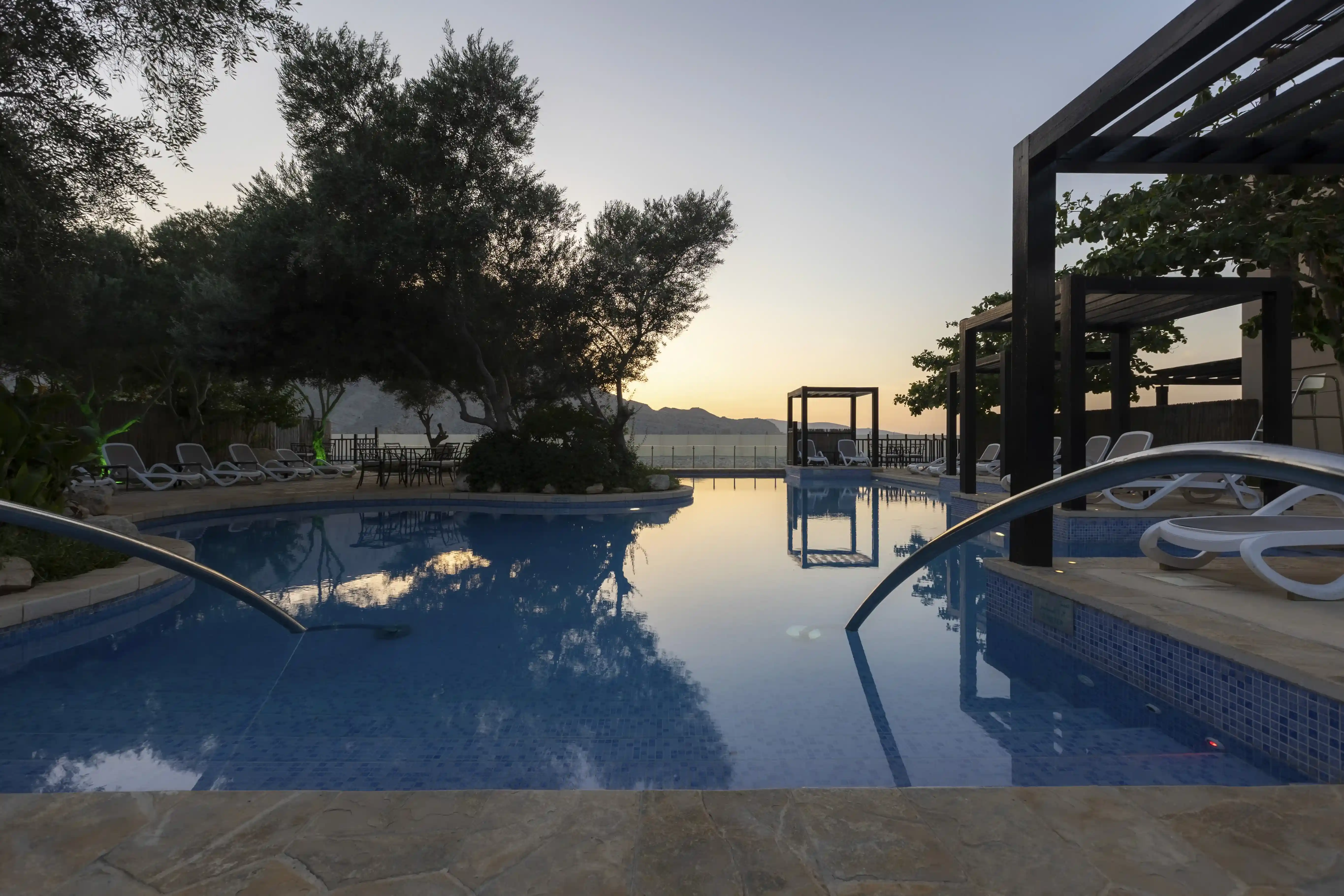 The main swimming pool at Atana Musandam at sunset, with the calm water reflecting the sky, poolside cabanas, and surrounding mountains.
