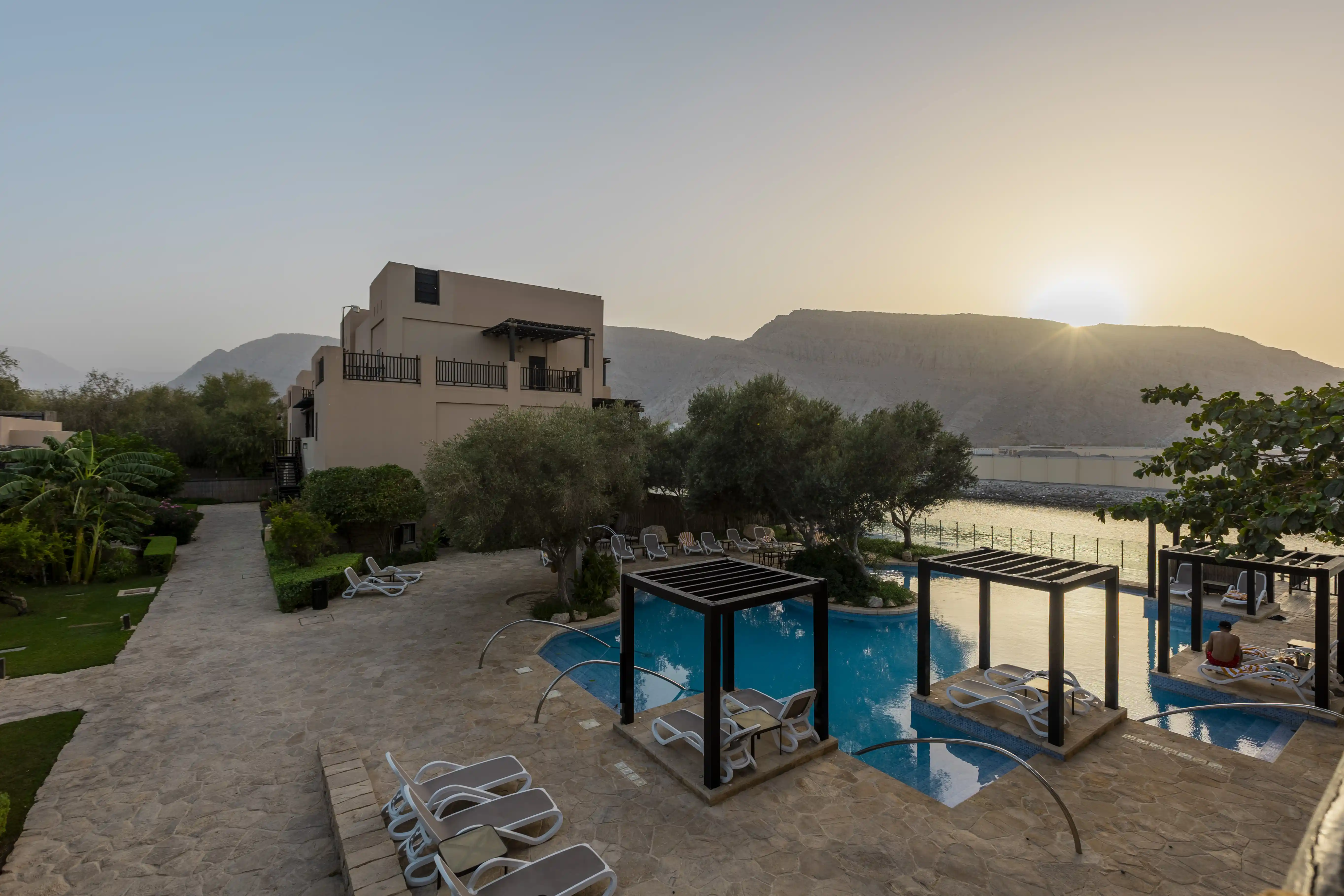 A top down view photo taken from a balcony at Atana Musandam of the swimming pool, at sun set. The sun peaks over the back edge of the mountains in the distance.