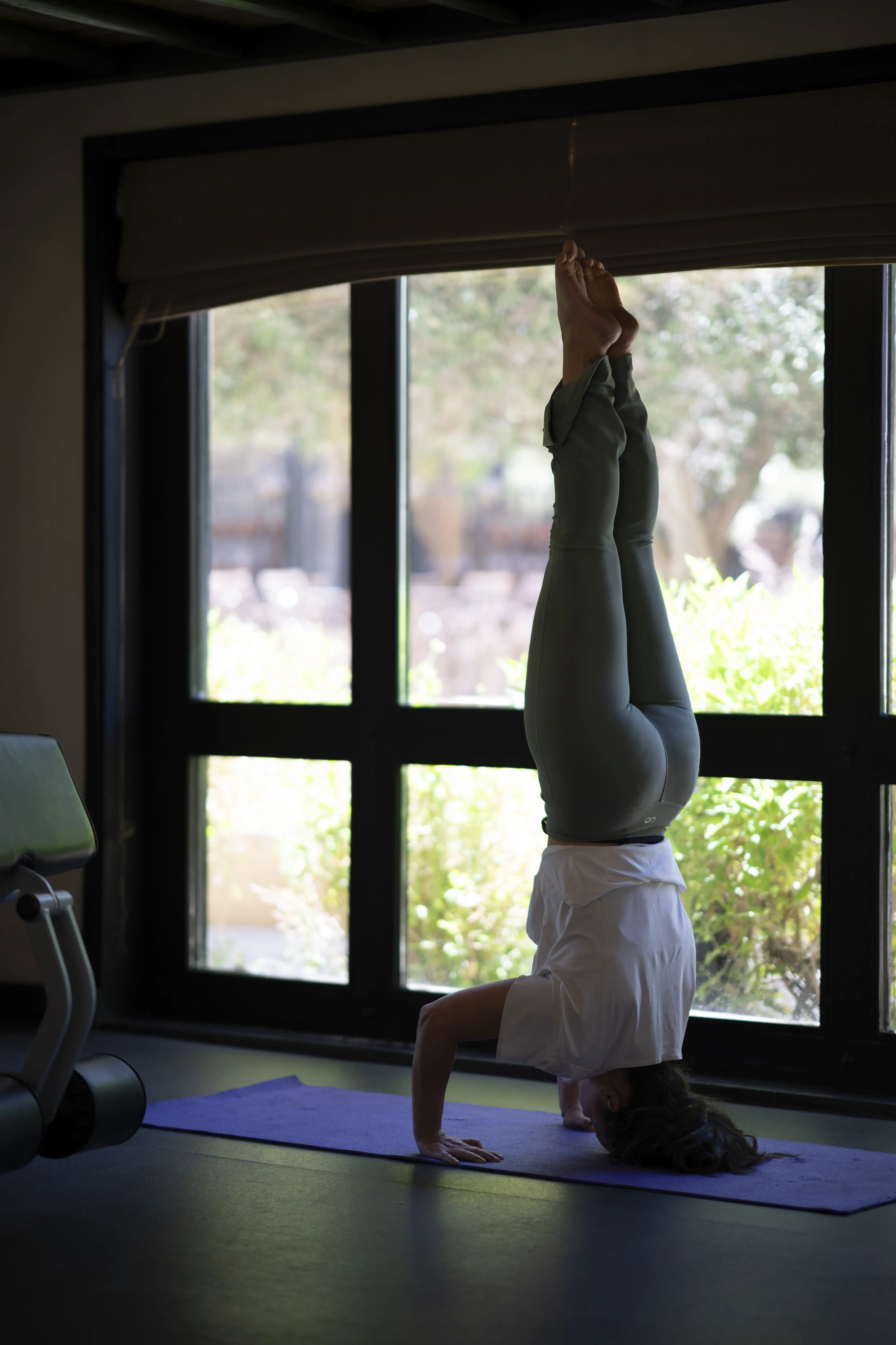 A woman practices a yoga headstand on a mat in the gym, silhouetted against a large window with a garden view.

