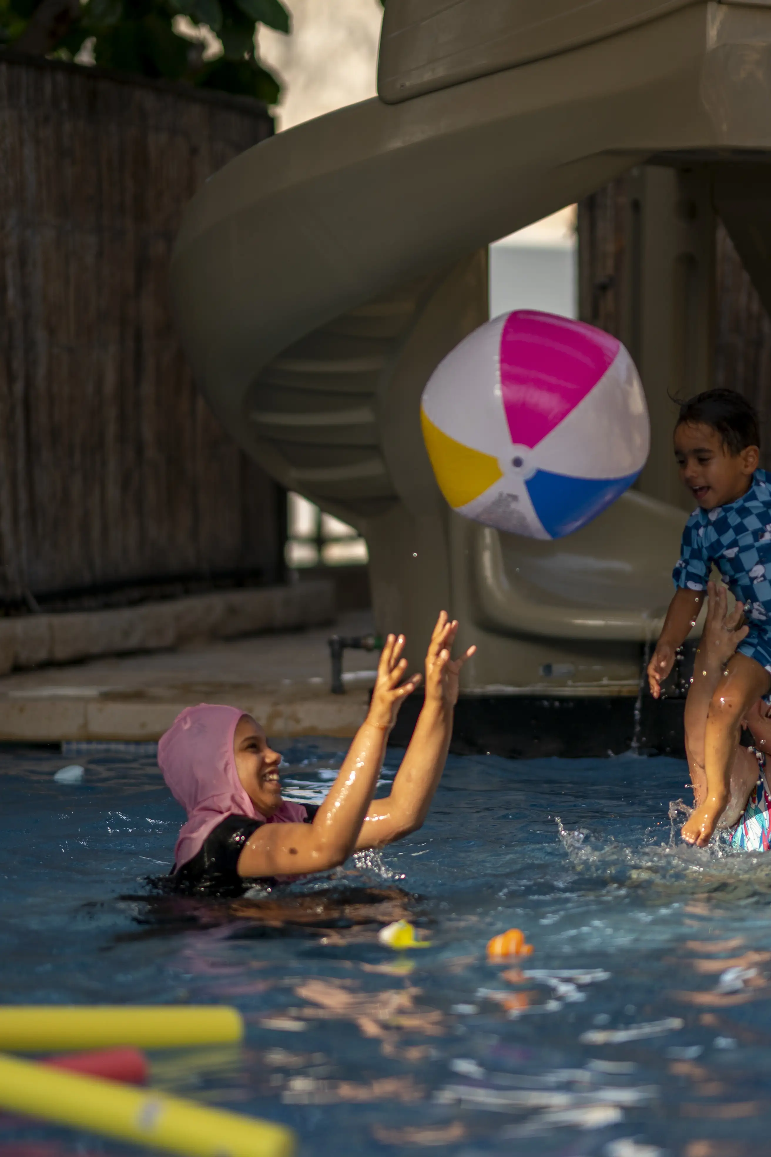 Two children have fun in the pool, laughing as they toss a large beach ball to each other.