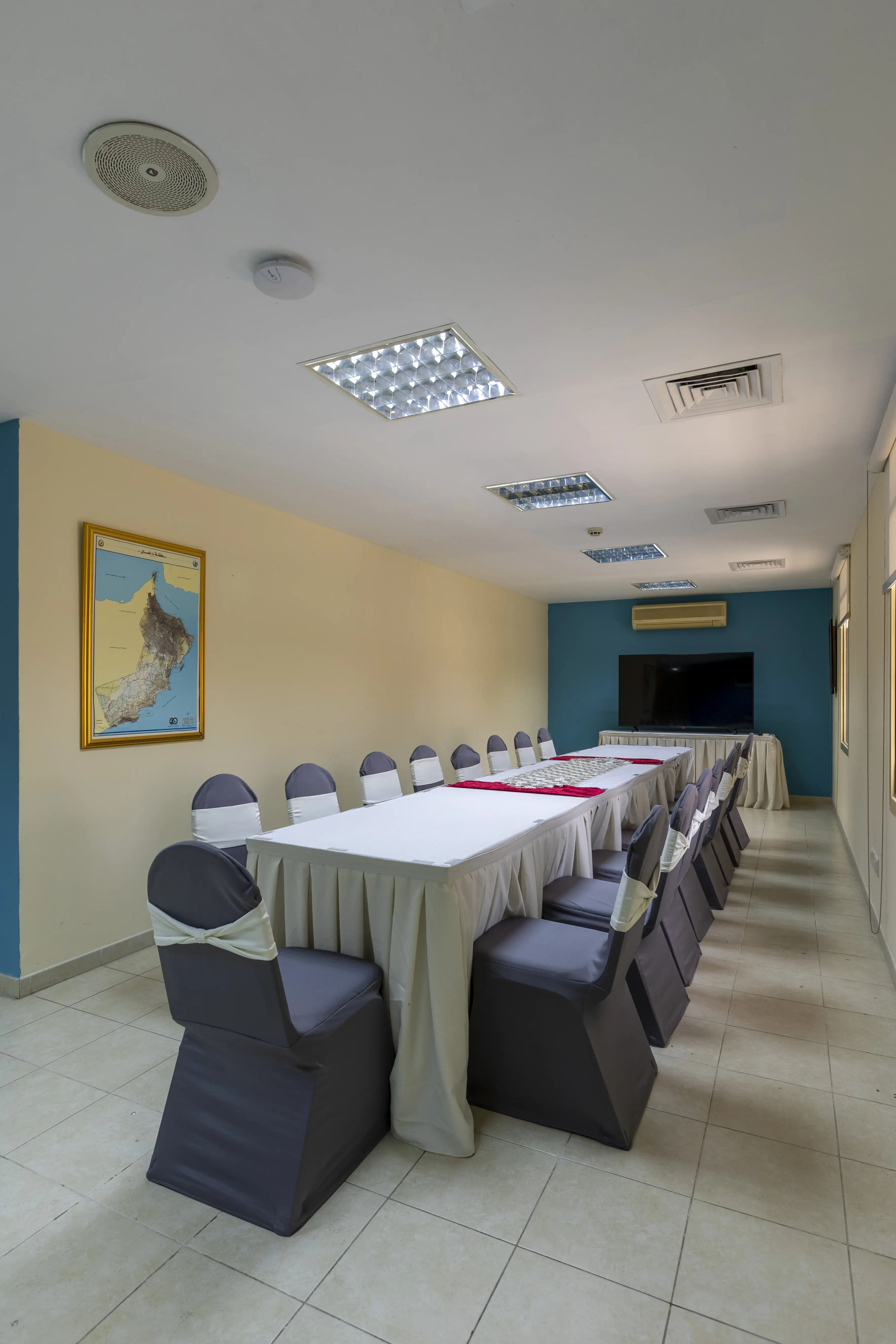 A long room with a long meeting room table, decorated with a table cloth. A blue wall with a tv at the back and chairs with chair covers around the table.