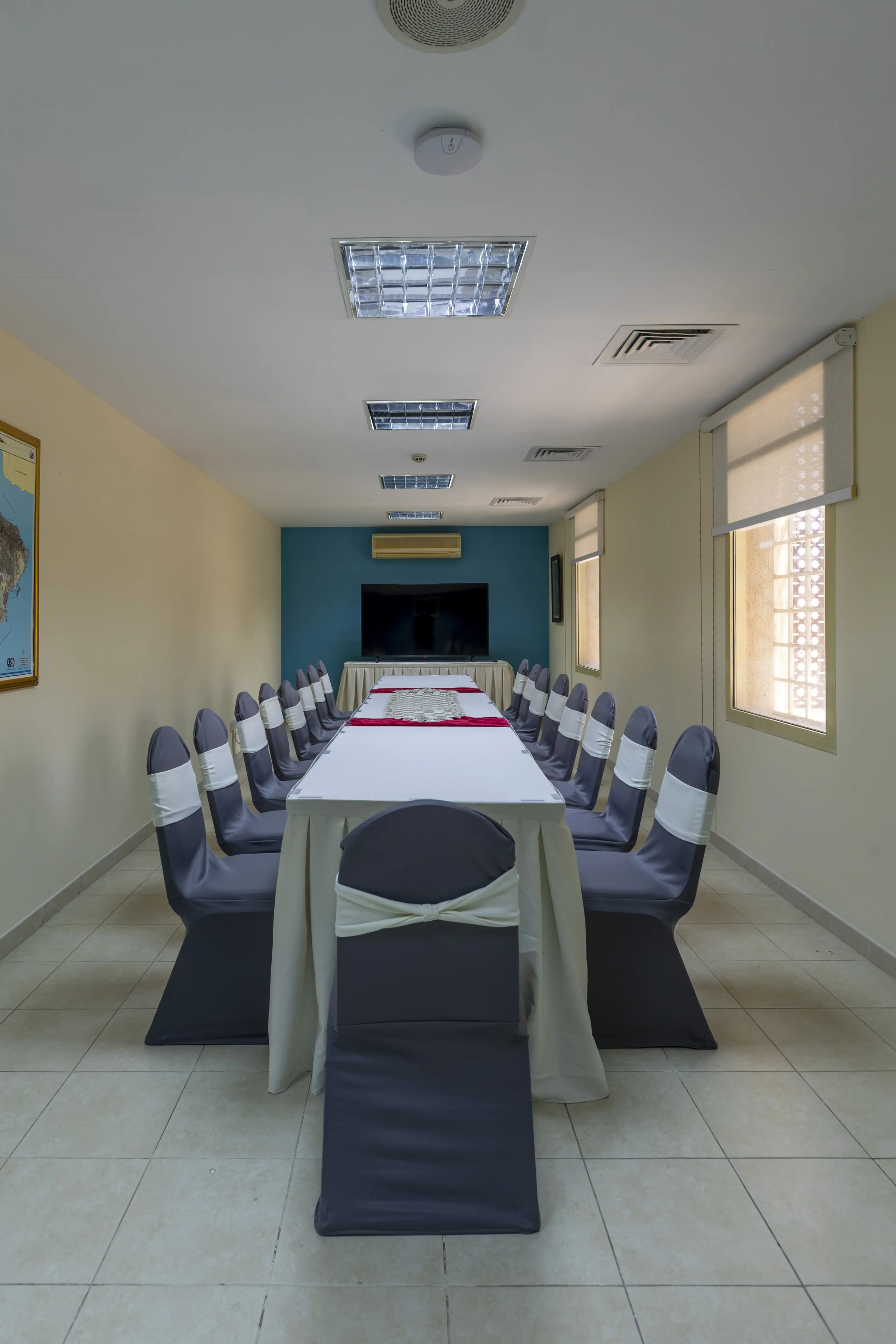 A long room with a long meeting room table, decorated with a table cloth. A blue wall with a tv at the back and chairs with chair covers around the table.