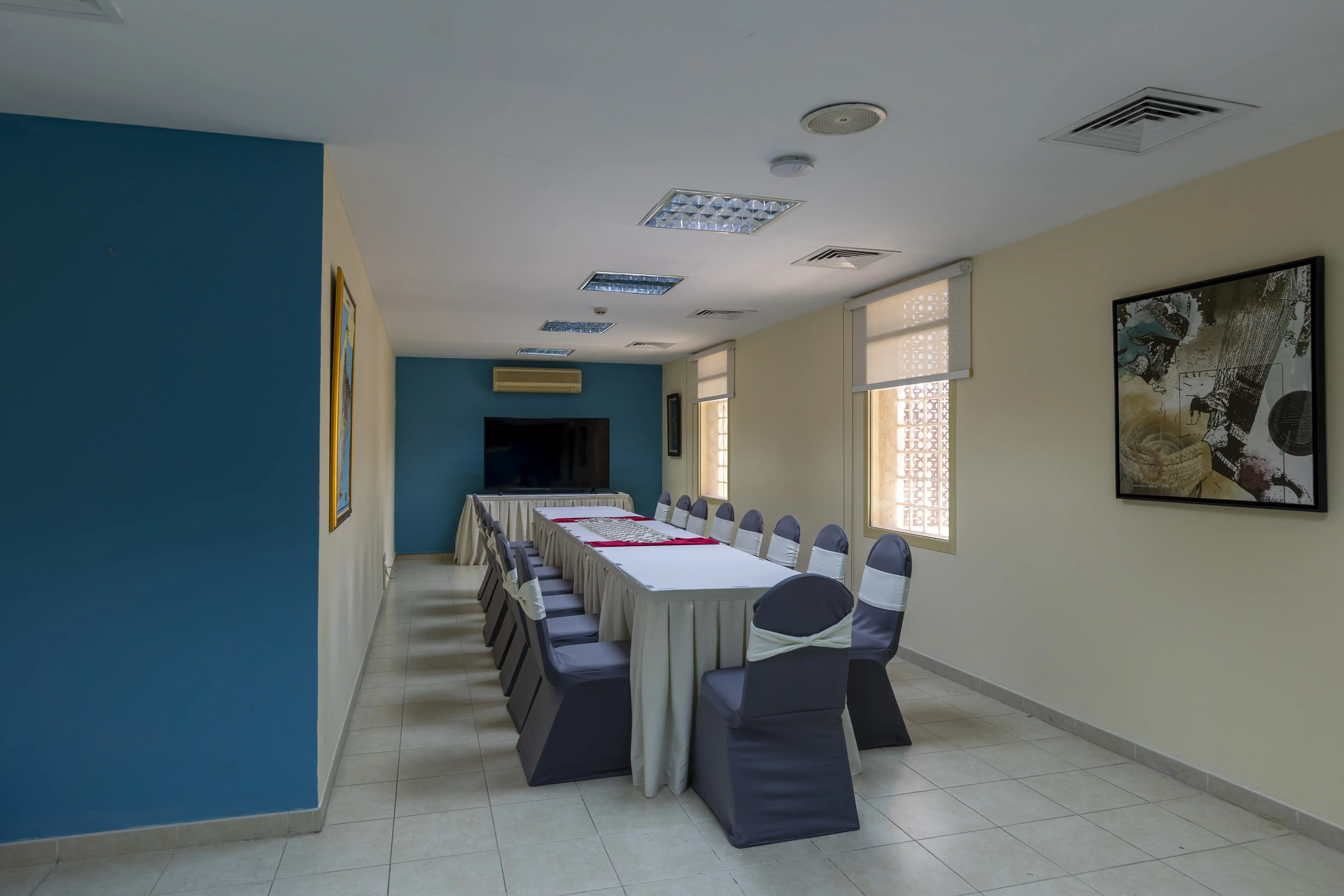 A long room with a long meeting room table, decorated with a table cloth. A blue wall with a tv at the back and chairs with chair covers around the table.