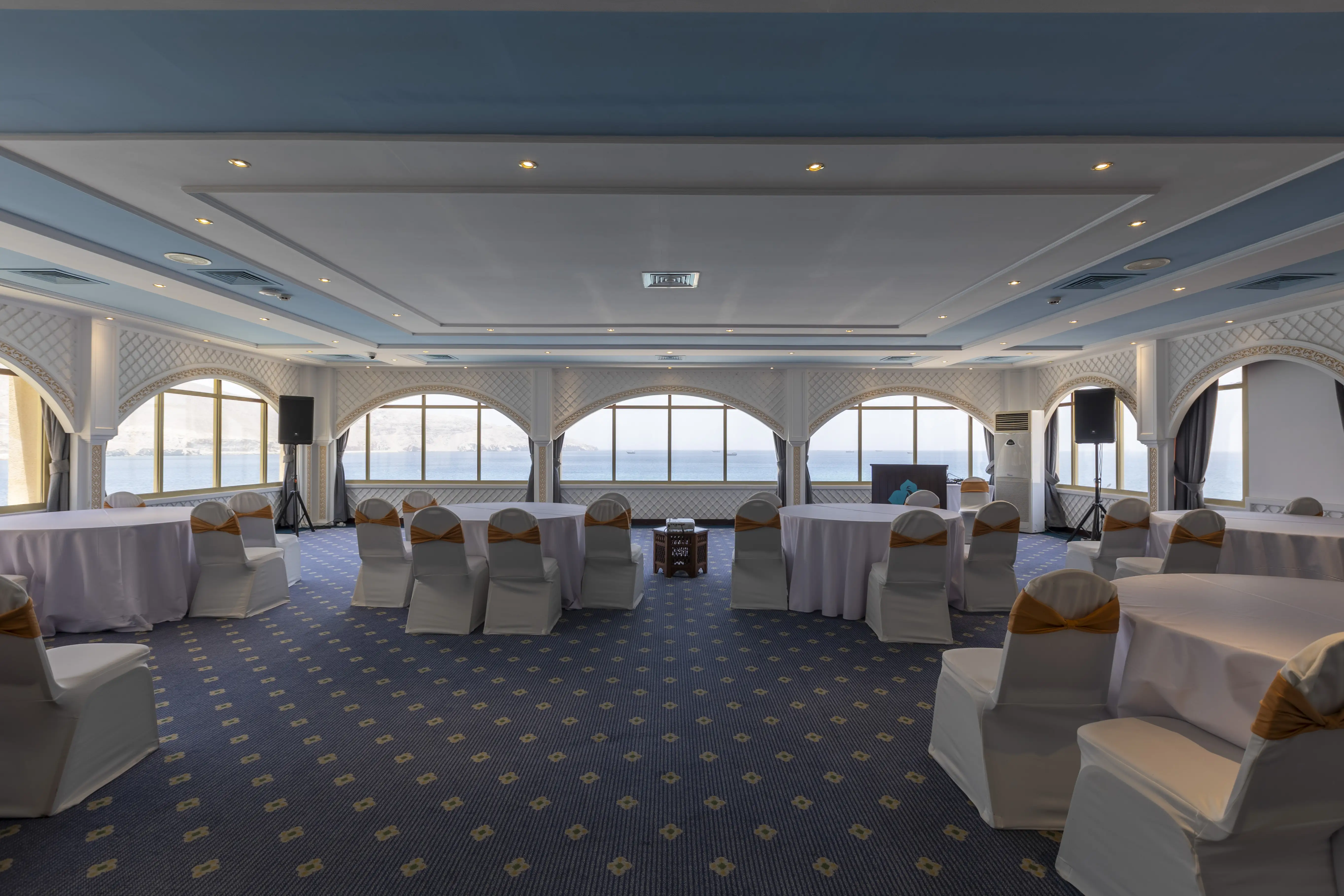 A large ball room with spectacular views of the ocean. Round tables with white table clothes scattered around the room, surrounded by banquet style chairs, with white seat covers. The ceiling as a distrinct baby blue/sky color. 