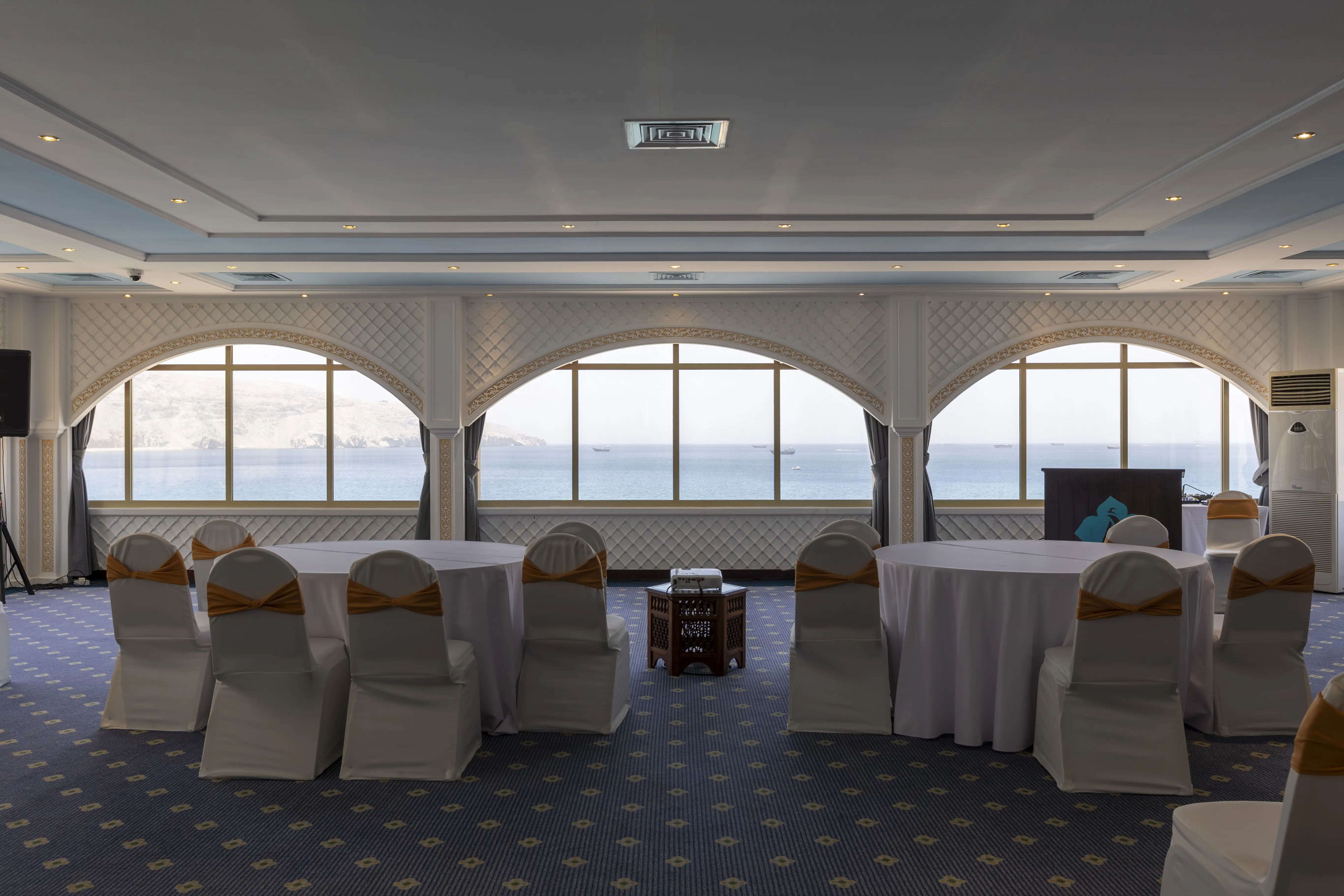 A large ball room with spectacular views of the ocean. Round tables with white table clothes scattered around the room, surrounded by banquet style chairs, with white seat covers. The ceiling as a distrinct baby blue/sky color. 