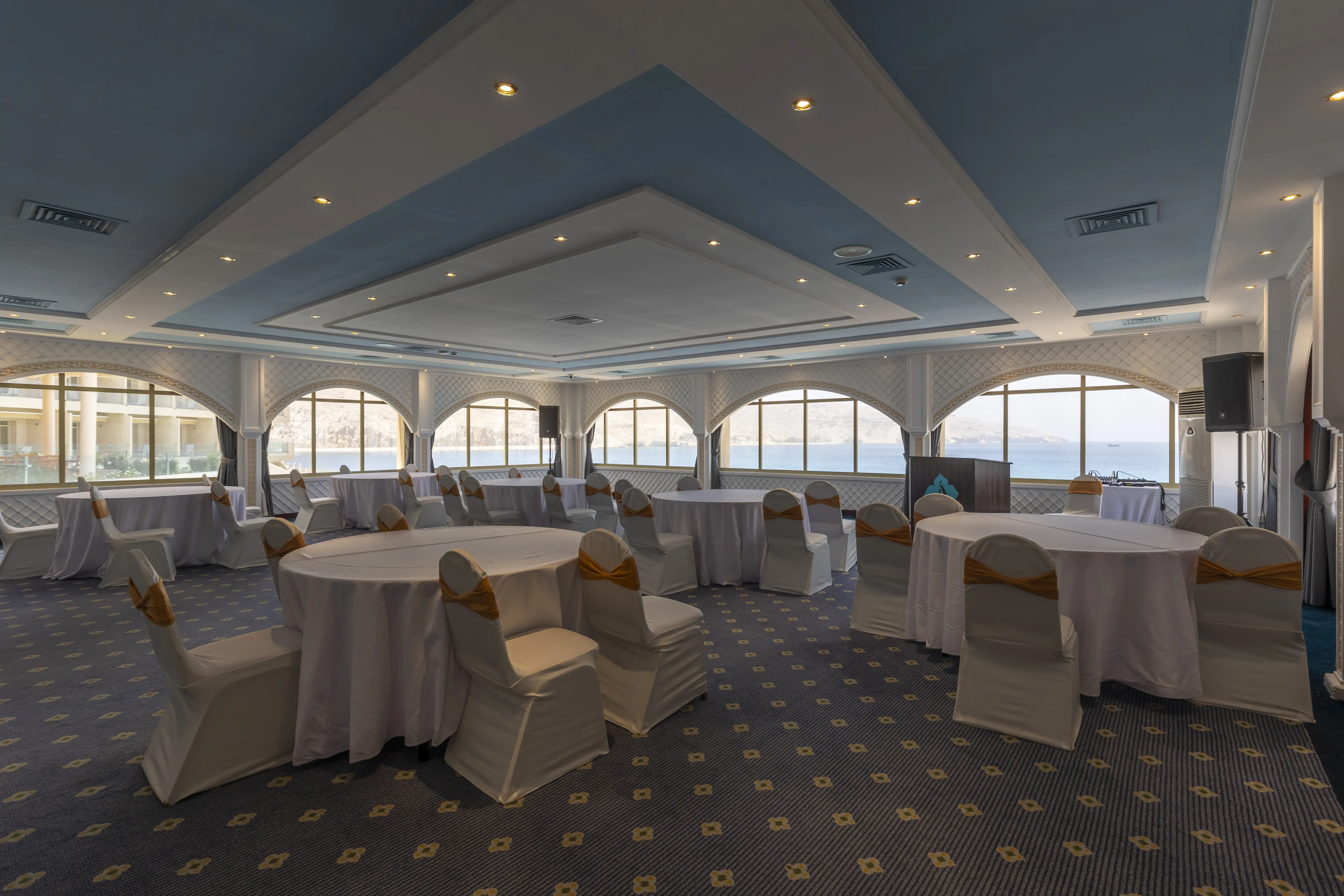 A large ball room with spectacular views of the ocean. Round tables with white table clothes scattered around the room, surrounded by banquet style chairs, with white seat covers. The ceiling as a distrinct baby blue/sky color. 