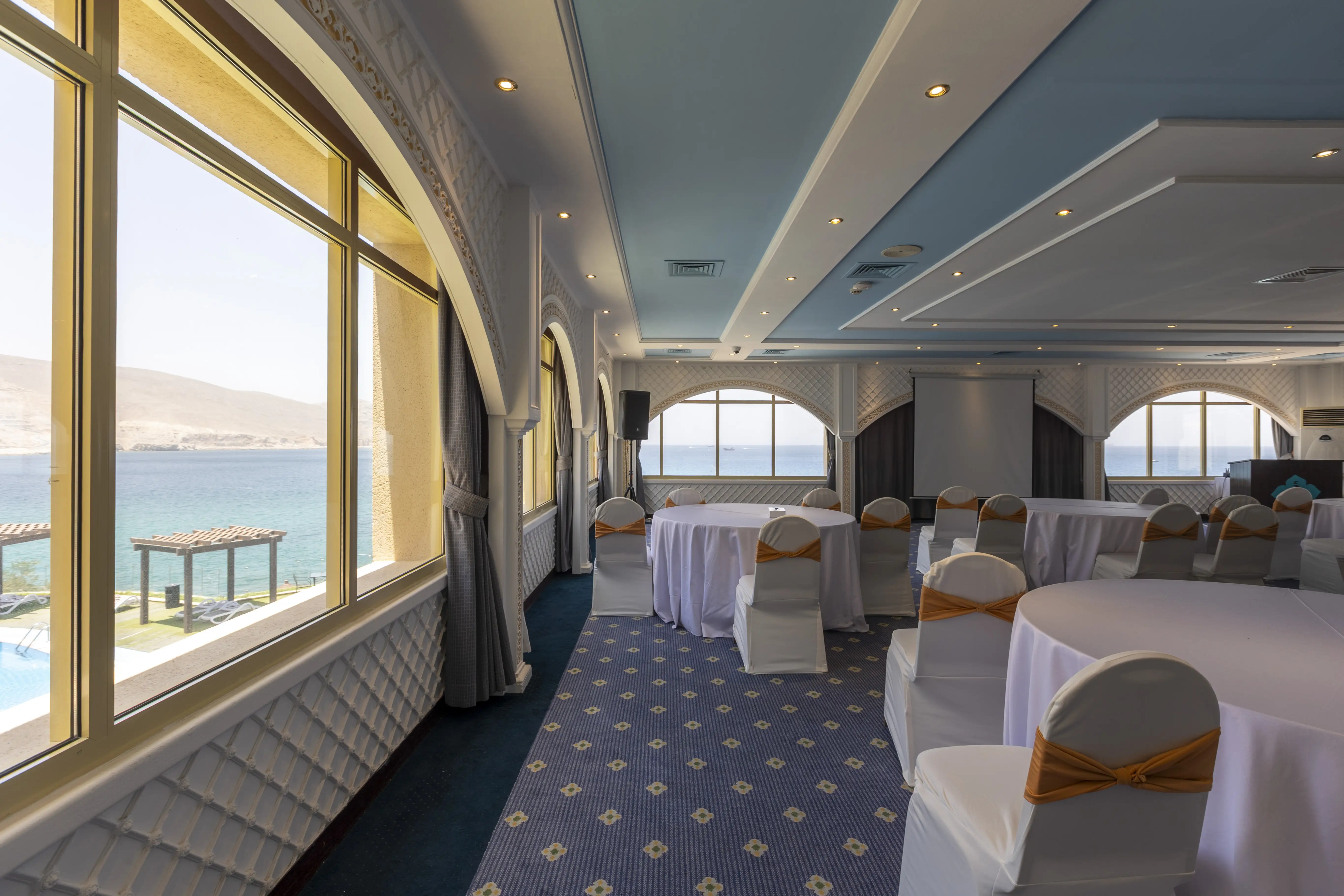 A large ball room with spectacular views of the ocean. Round tables with white table clothes scattered around the room, surrounded by banquet style chairs, with white seat covers. The ceiling as a distrinct baby blue/sky color. 