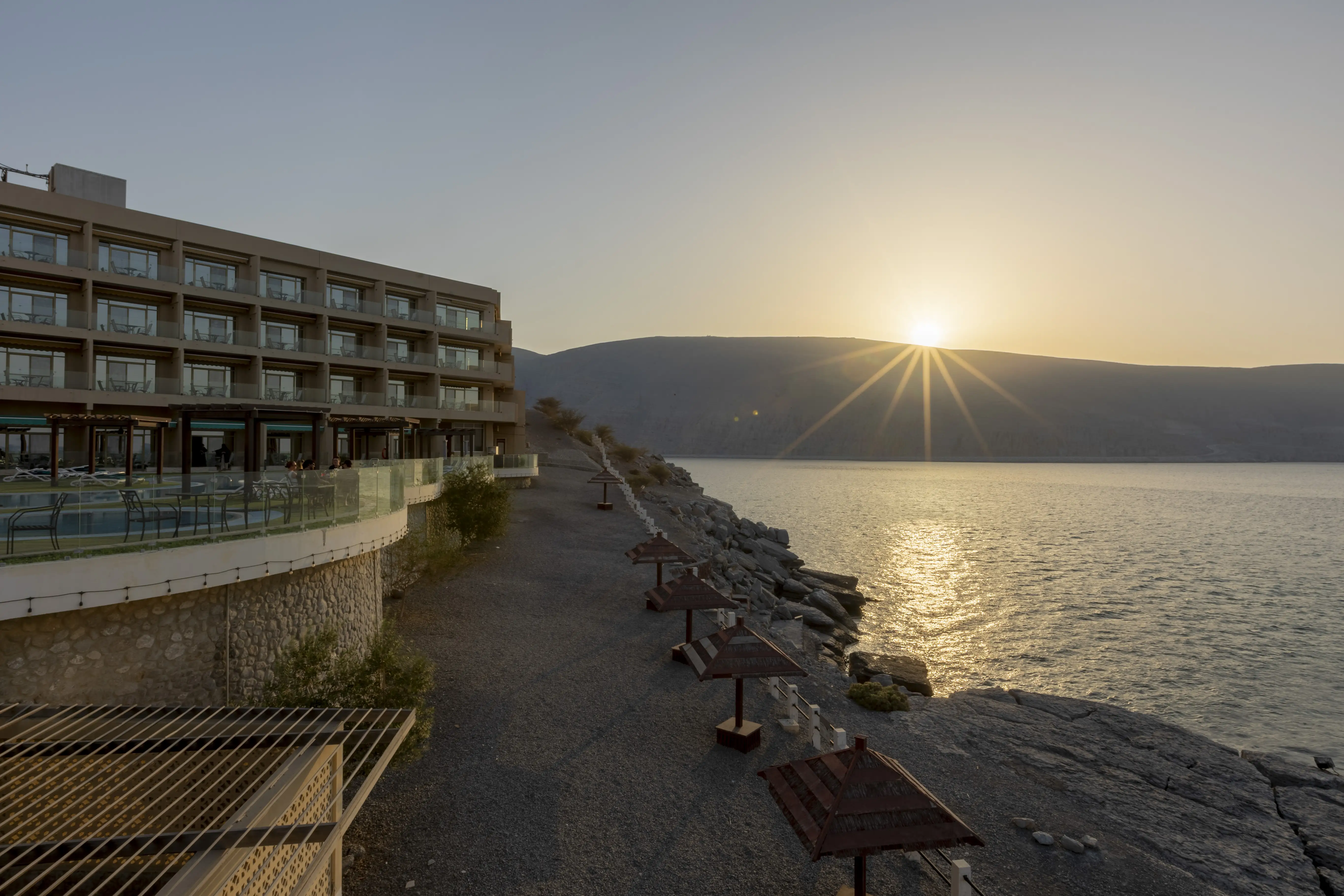 Sunset in the distance, light reflected on the pool at Atana Khasab, and showing the cliff edges and lower section of the property. With ocean on the right and the hotel on the left.