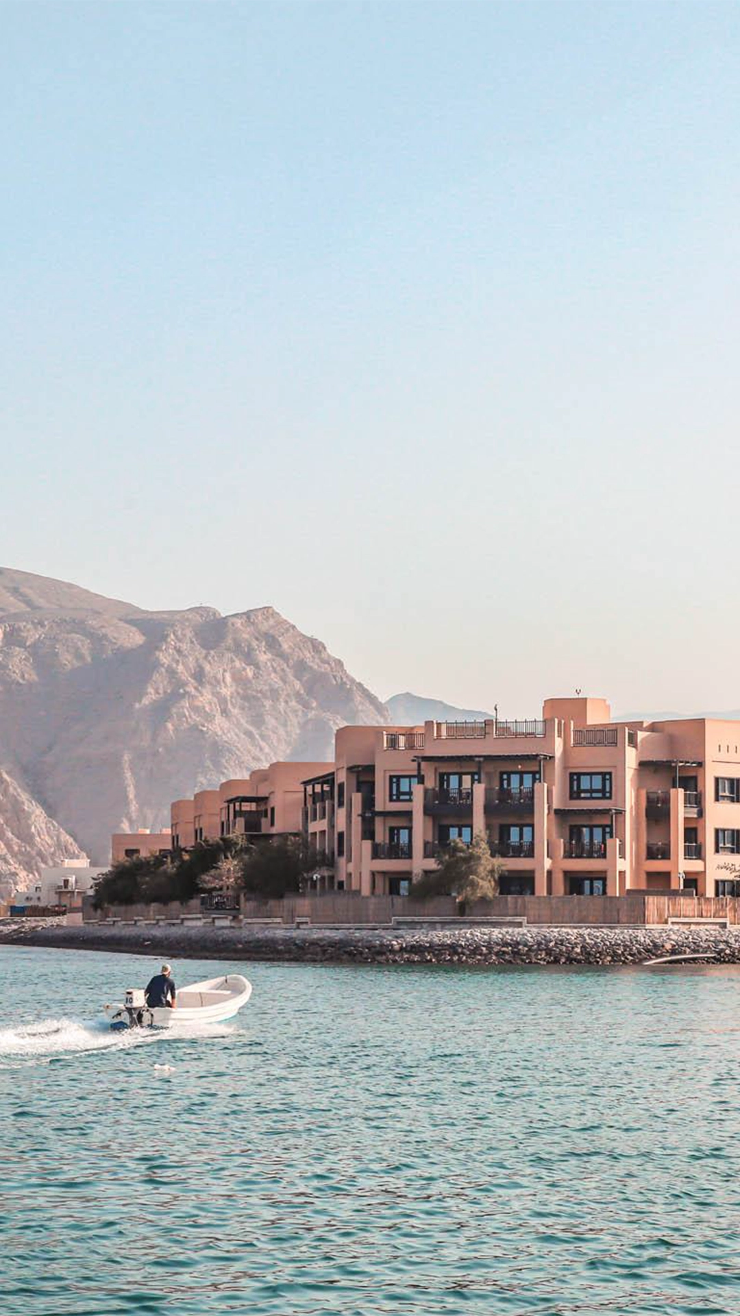 Atana Musandam property image taken from the water with the mountains in the background. A small speedboat rides in from the left of the image.