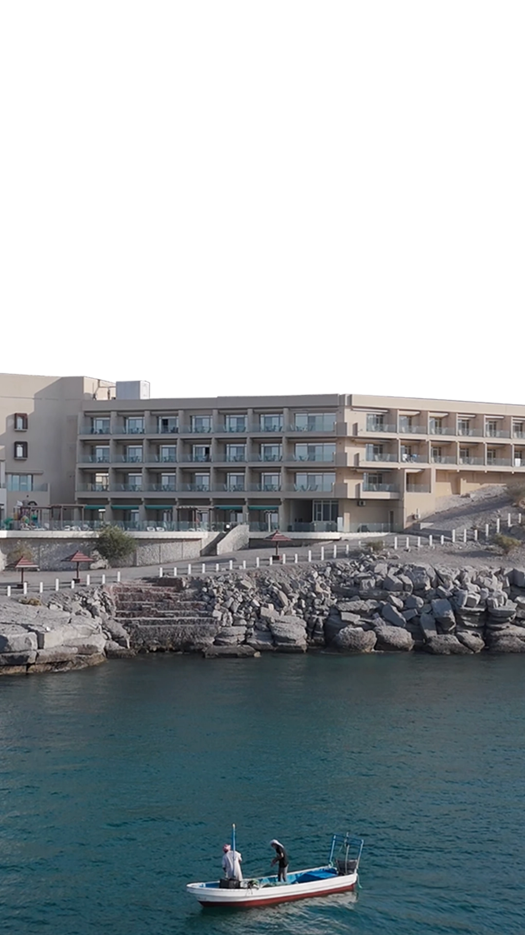Atana Khasab hotel taken from an aerial perspective, out in the water. In the foreground, there is water with 2 men standing on a small fishing boat.