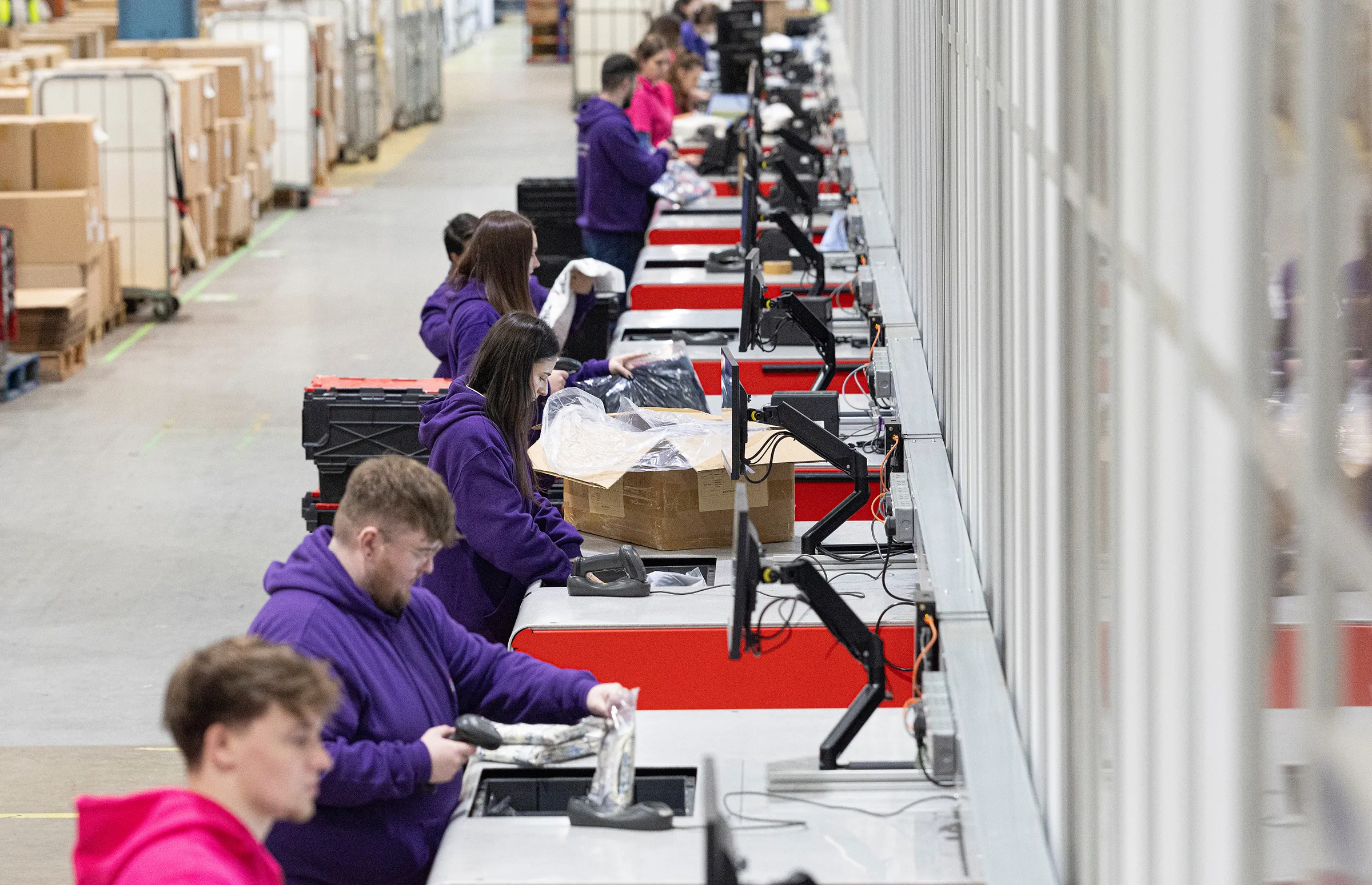 Workers in purple hoodies scanning and packing items on a long warehouse conveyor line.