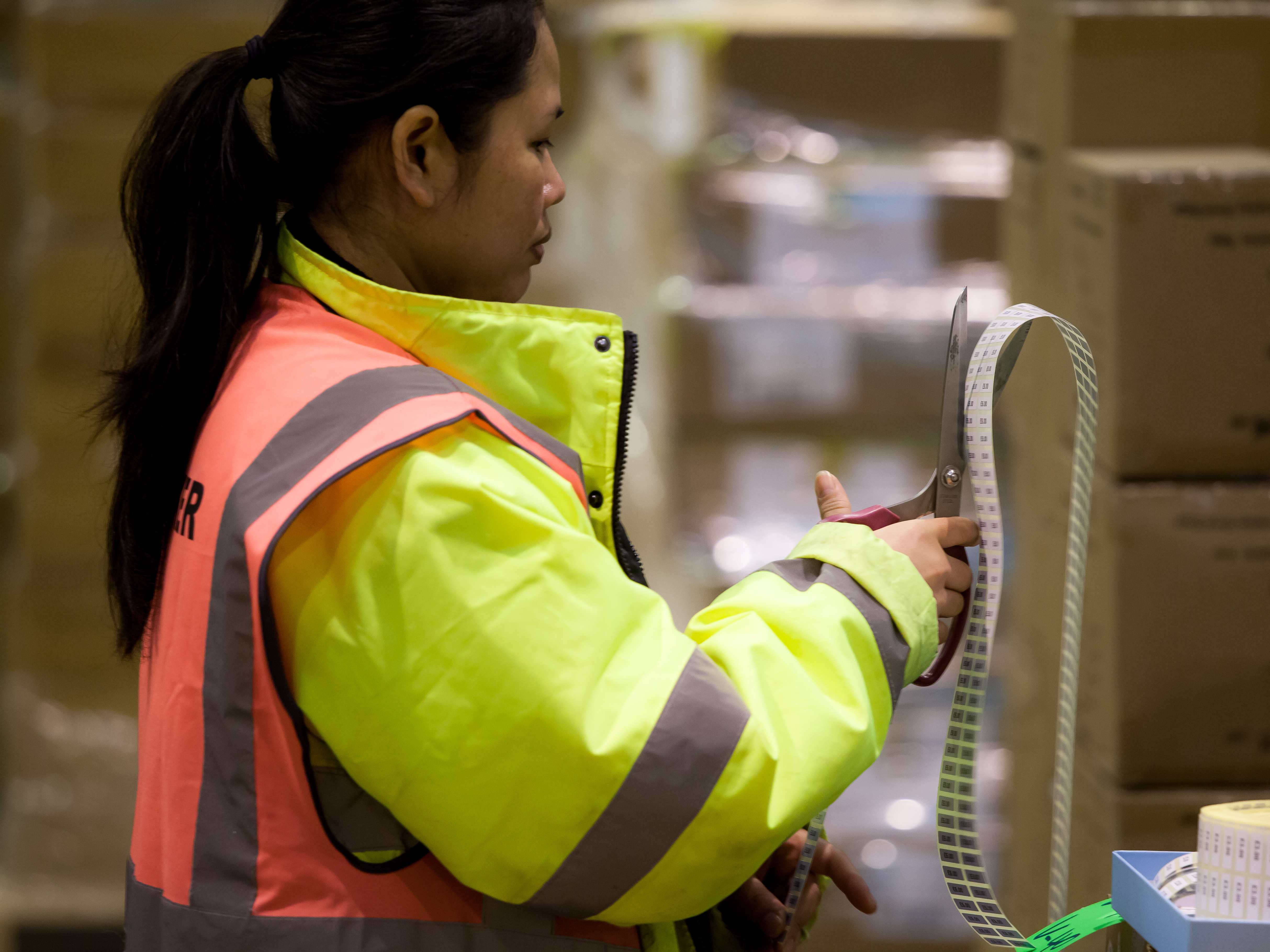 Woman in high-visibility jacket cutting a strip of barcode labels with scissors in a warehouse.