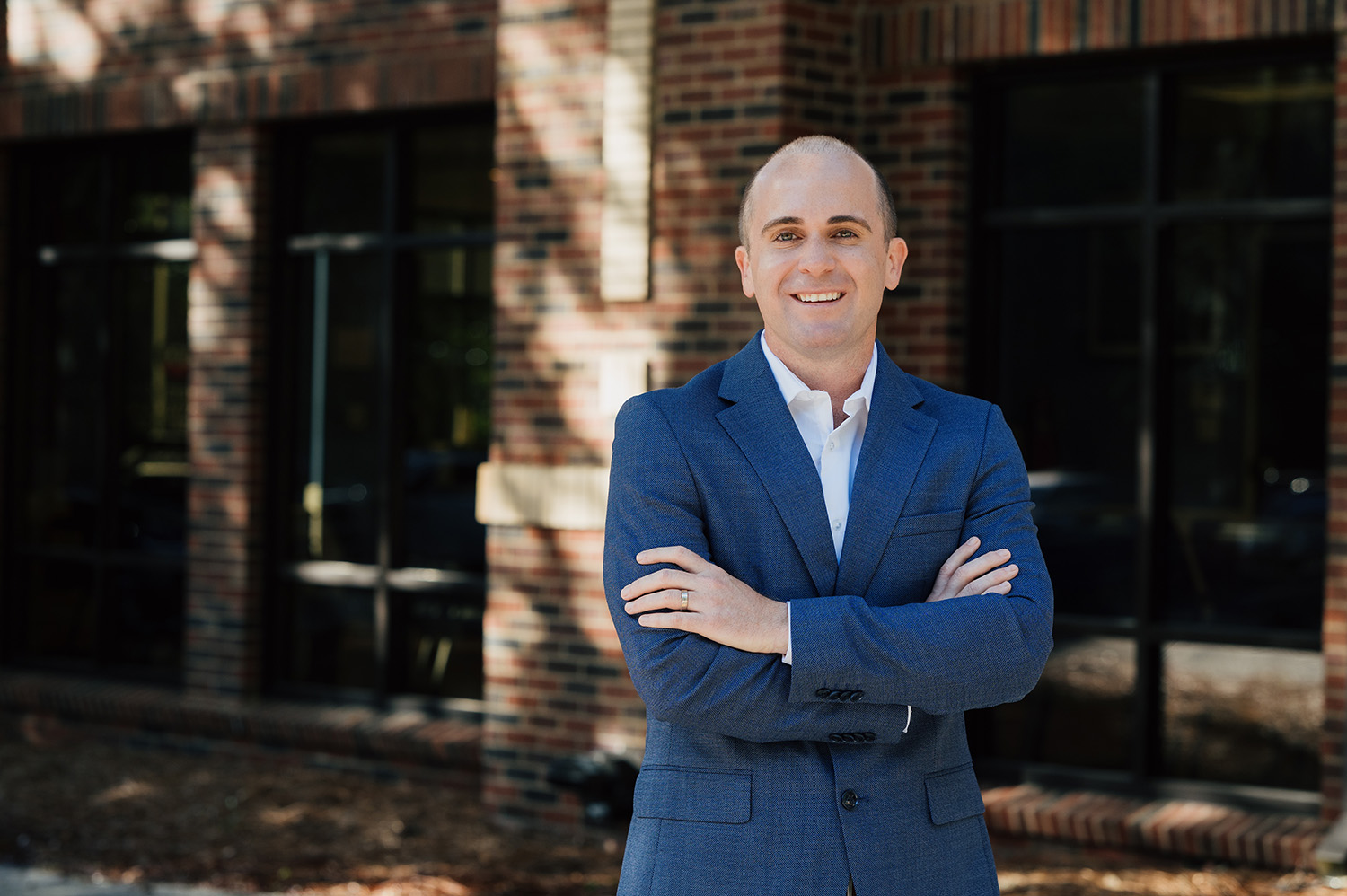 Smiling man in a blue suit with arms crossed standing outside a brick building.