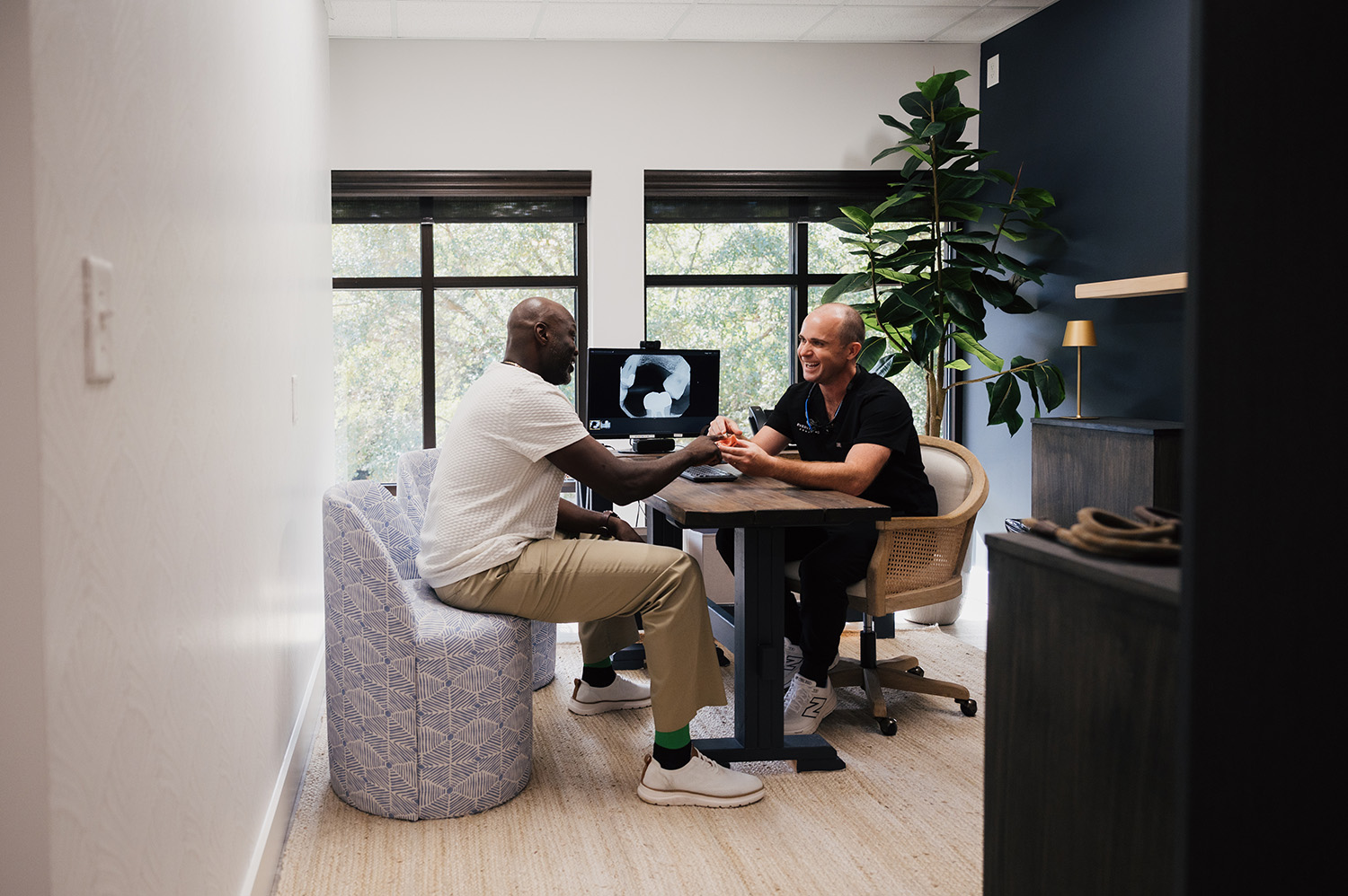 Two men seated in a modern office discussing a dental model with a dental X-ray displayed on a computer screen behind them.