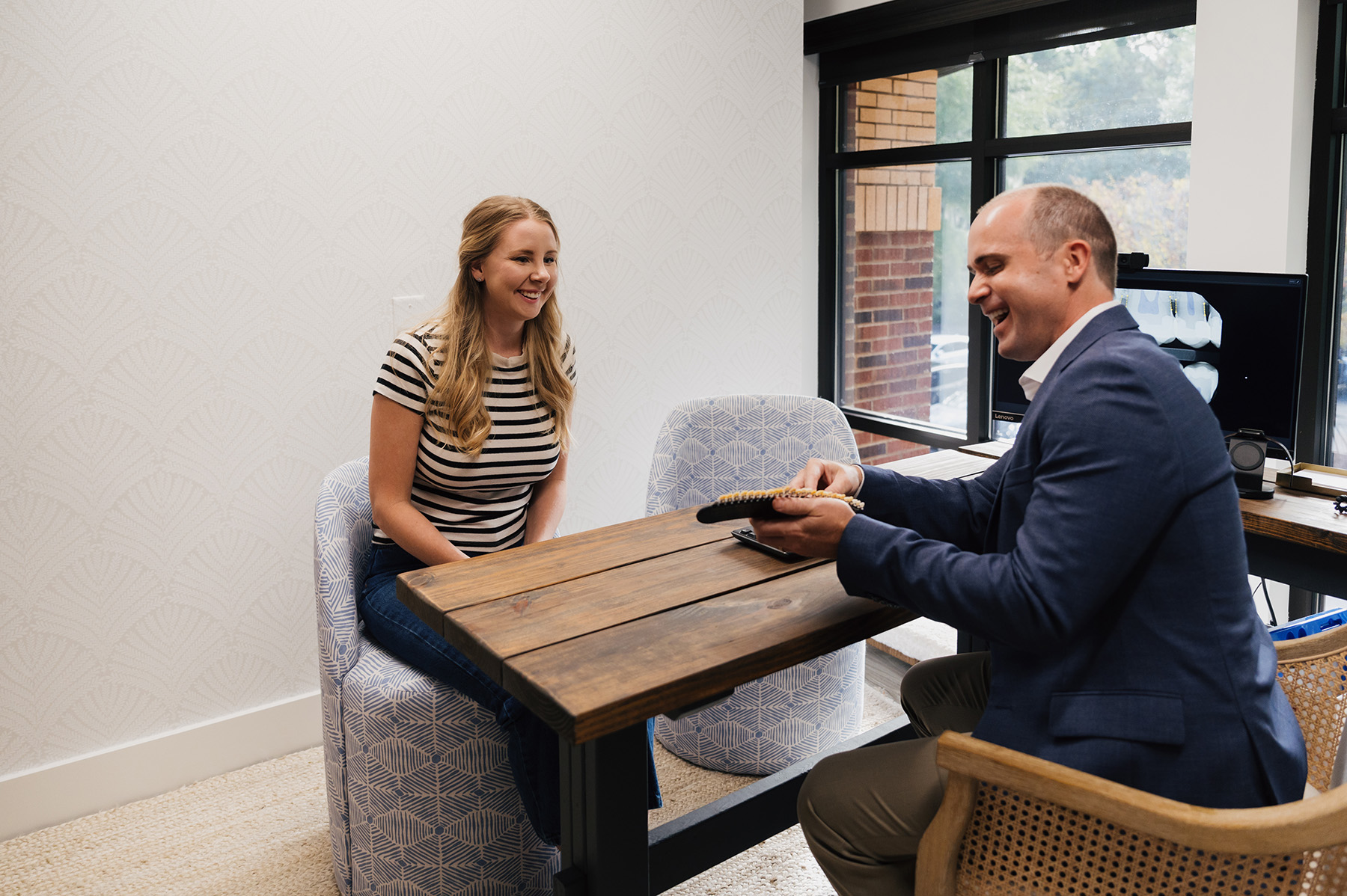 Woman in striped shirt smiling while sitting across a wooden table from a man in a blazer holding a notebook in a bright office.