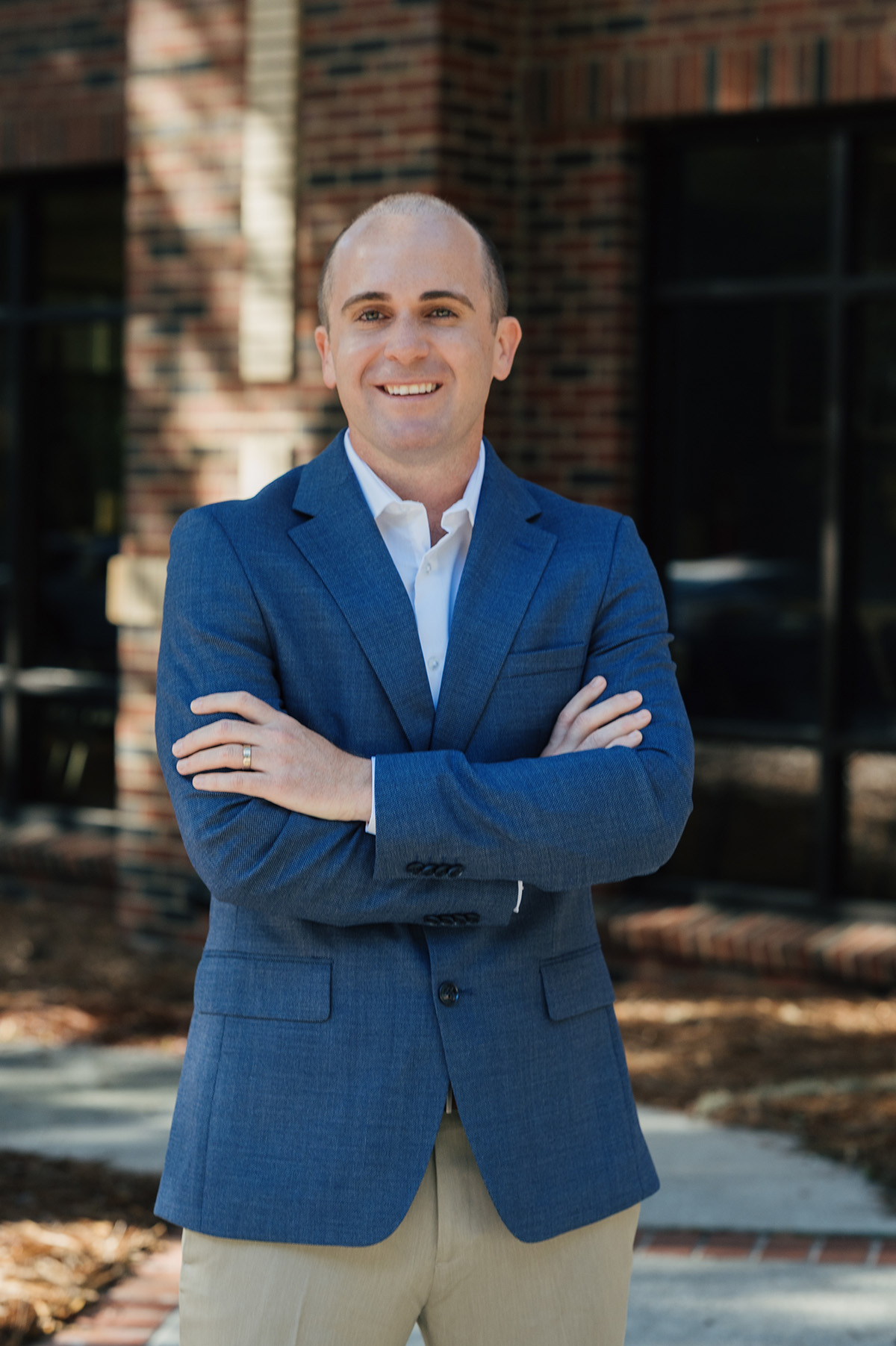 Smiling man in blue blazer and khaki pants standing with arms crossed outside a brick building.