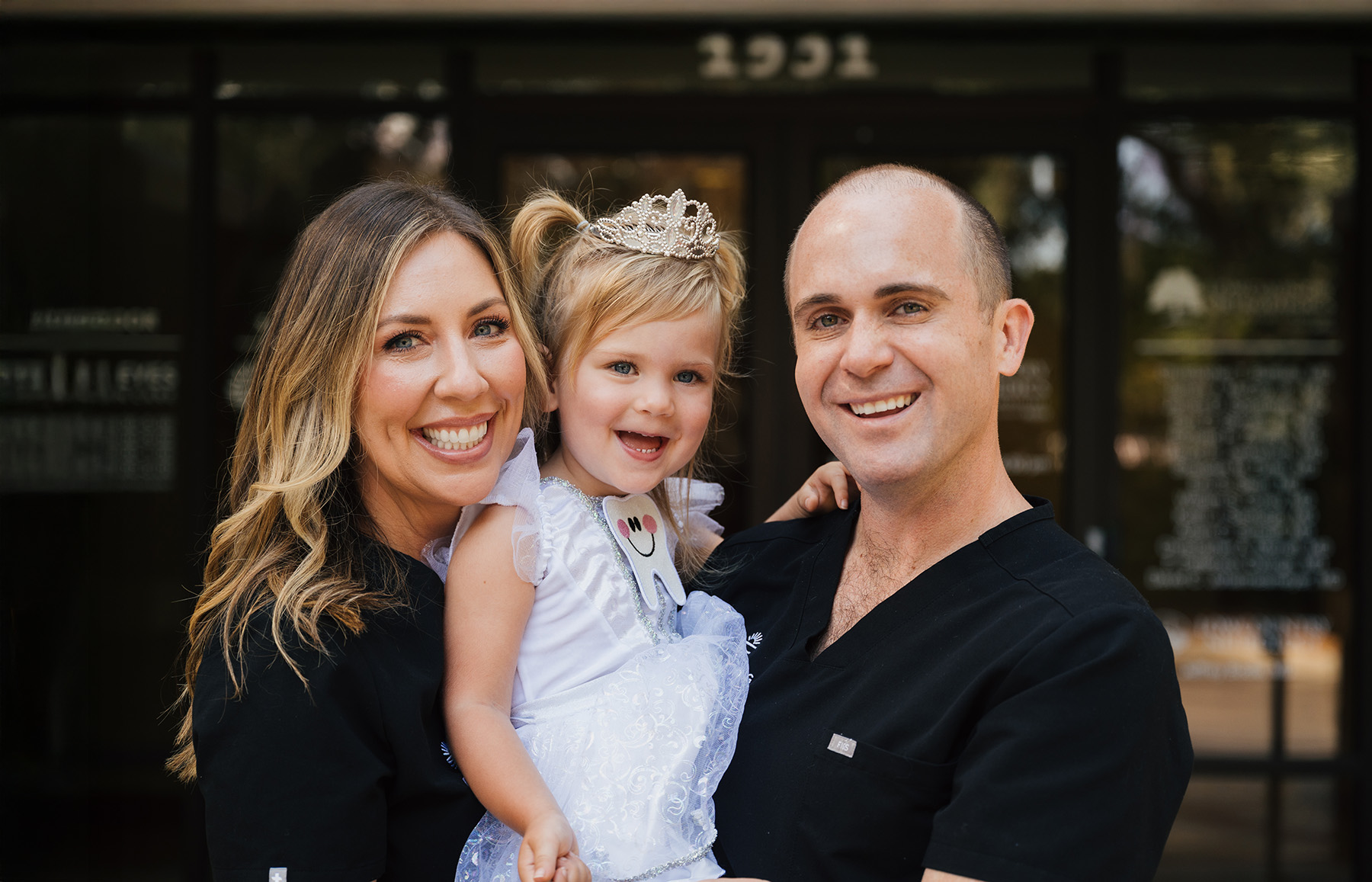 Smiling man and woman in black scrubs holding a young girl dressed as a princess with a tooth emblem.
