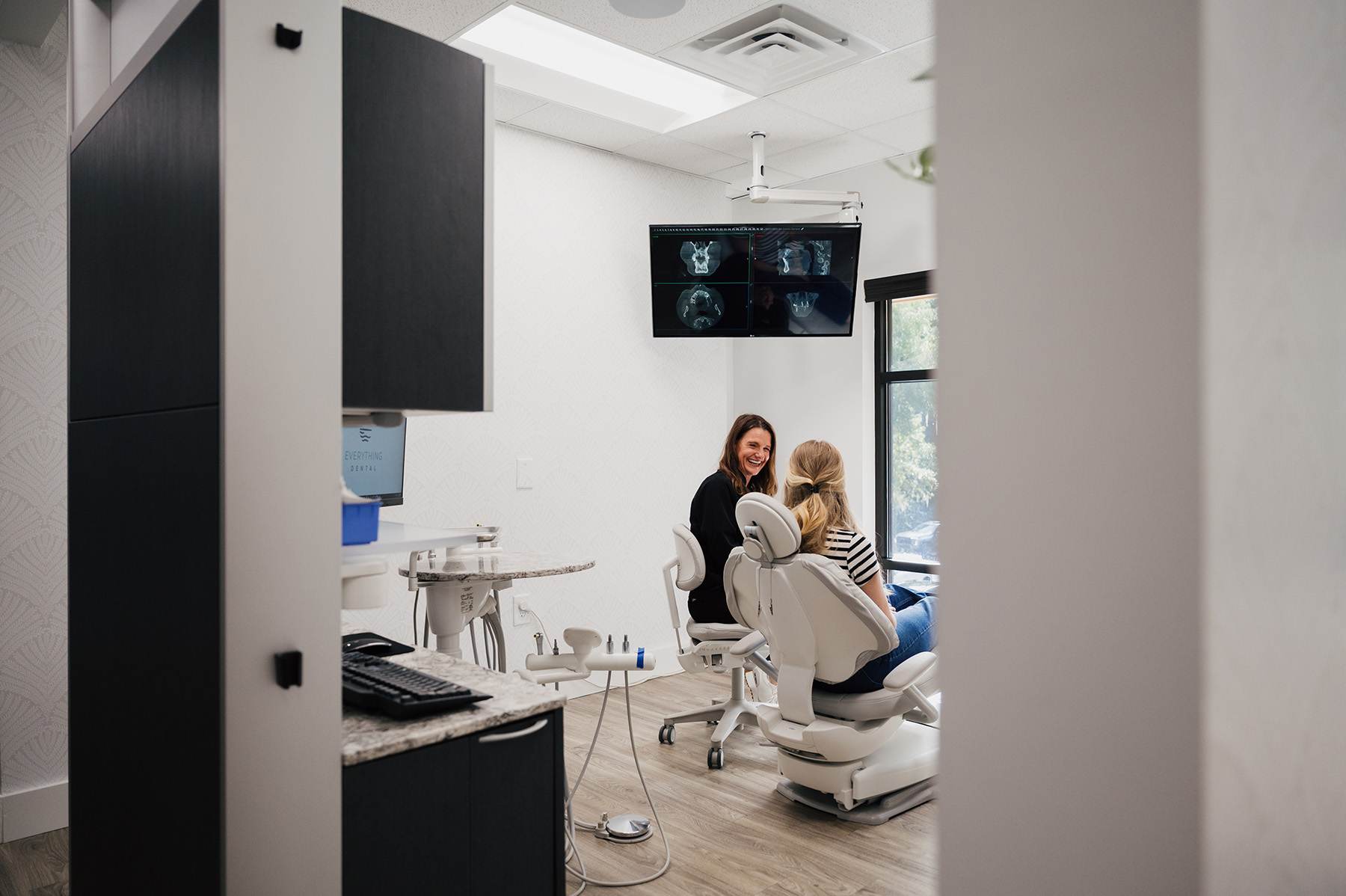 Dentist smiling and talking with a patient seated in a dental chair in a modern clinic room showing dental X-rays on a screen.