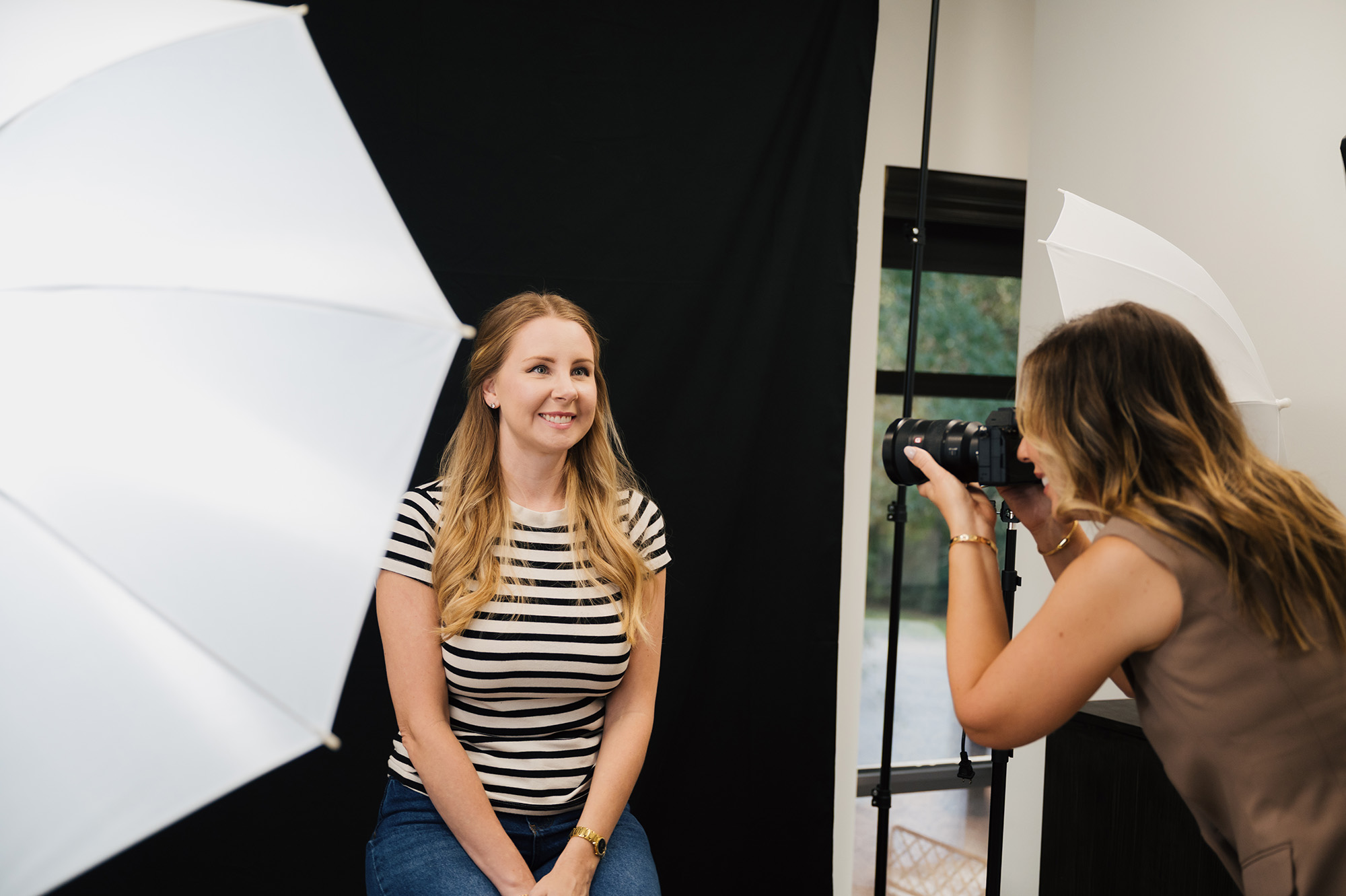 Photographer taking a photo of a smiling woman sitting against a black backdrop in a studio with umbrella lights.