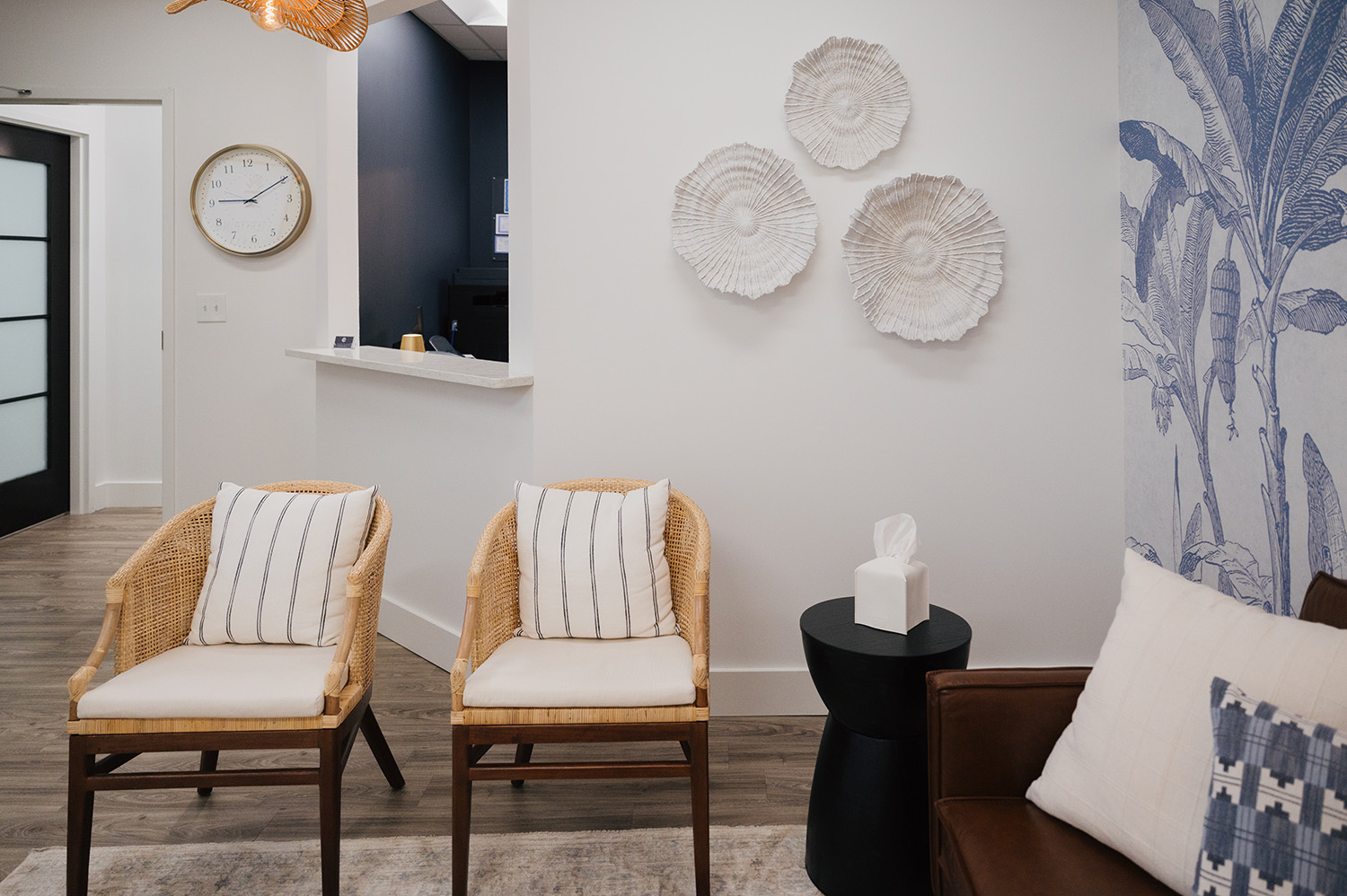 Modern waiting room with two wicker chairs with striped pillows, a black side table with a tissue box, decorative wall art, and a brown leather couch with pillows.