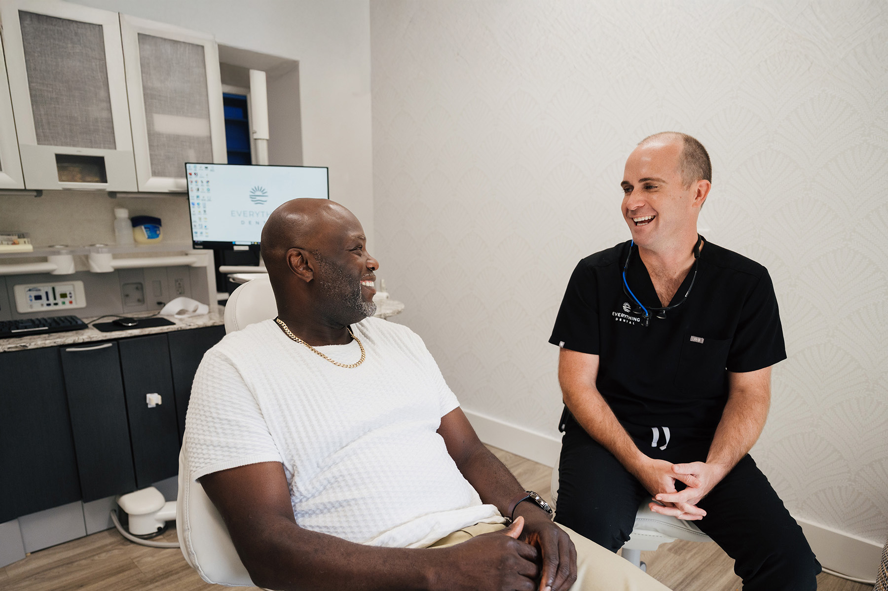 A male dentist in black scrubs smiling and talking with a male patient seated in a dental chair.