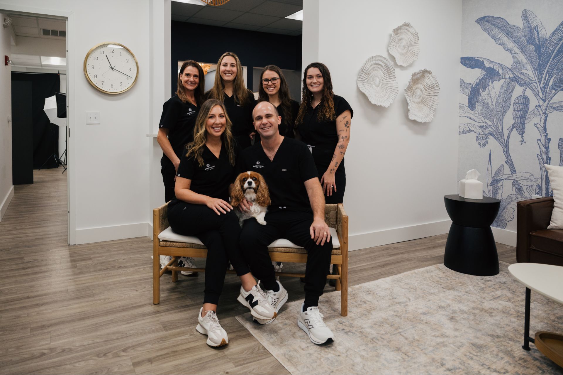 Six dental professionals dressed in black scrubs posing indoors with a Cavalier King Charles Spaniel dog, sitting on a wooden bench in a modern office with light wood flooring and blue botanical wall art.