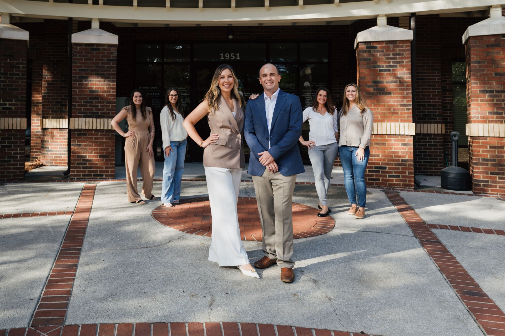Group of six people smiling and posing outside a brick building with the number 1951 above the entrance.