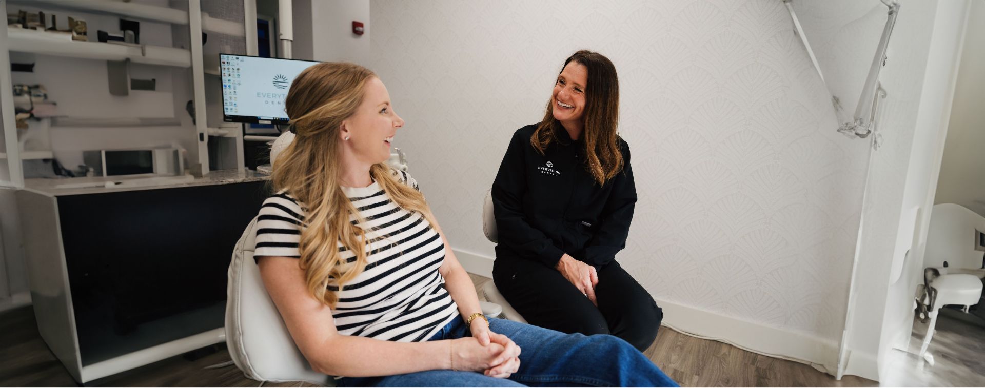 Two women smiling and talking in a dental office, one seated in a patient chair and the other in a chair wearing dental scrubs.