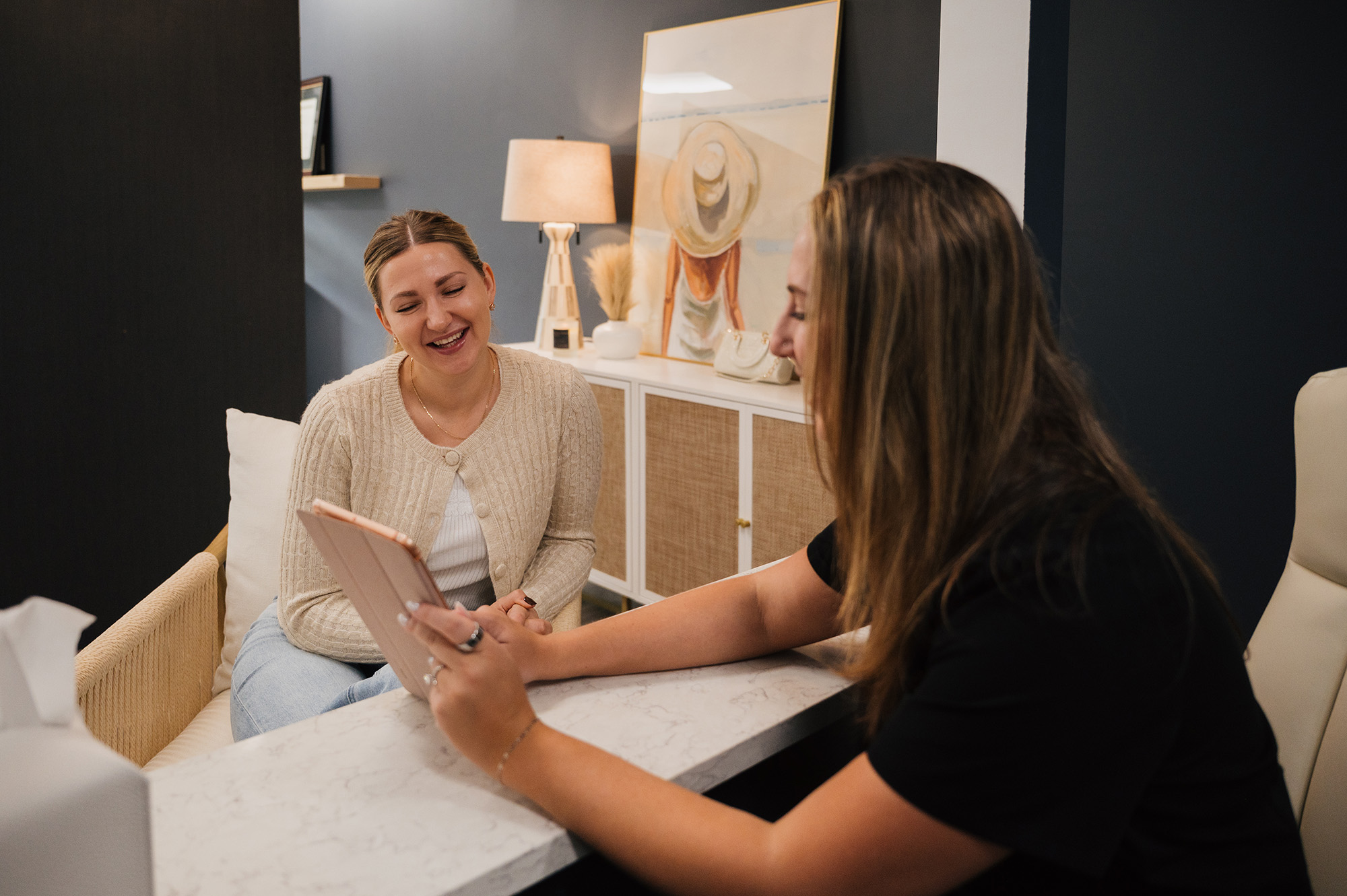 Two women sitting across a marble desk, one showing the other something on a tablet, both smiling in a cozy office setting.