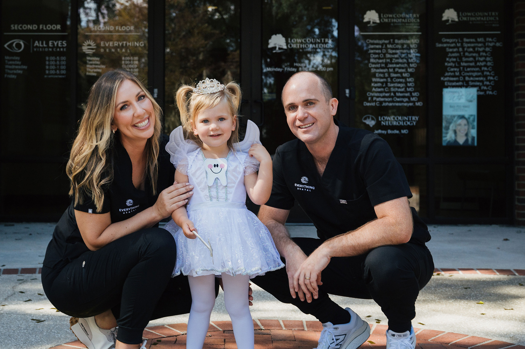 Smiling woman and man in black scrubs posing outside a dental office with a young girl dressed as a tooth fairy in a white dress and tiara.