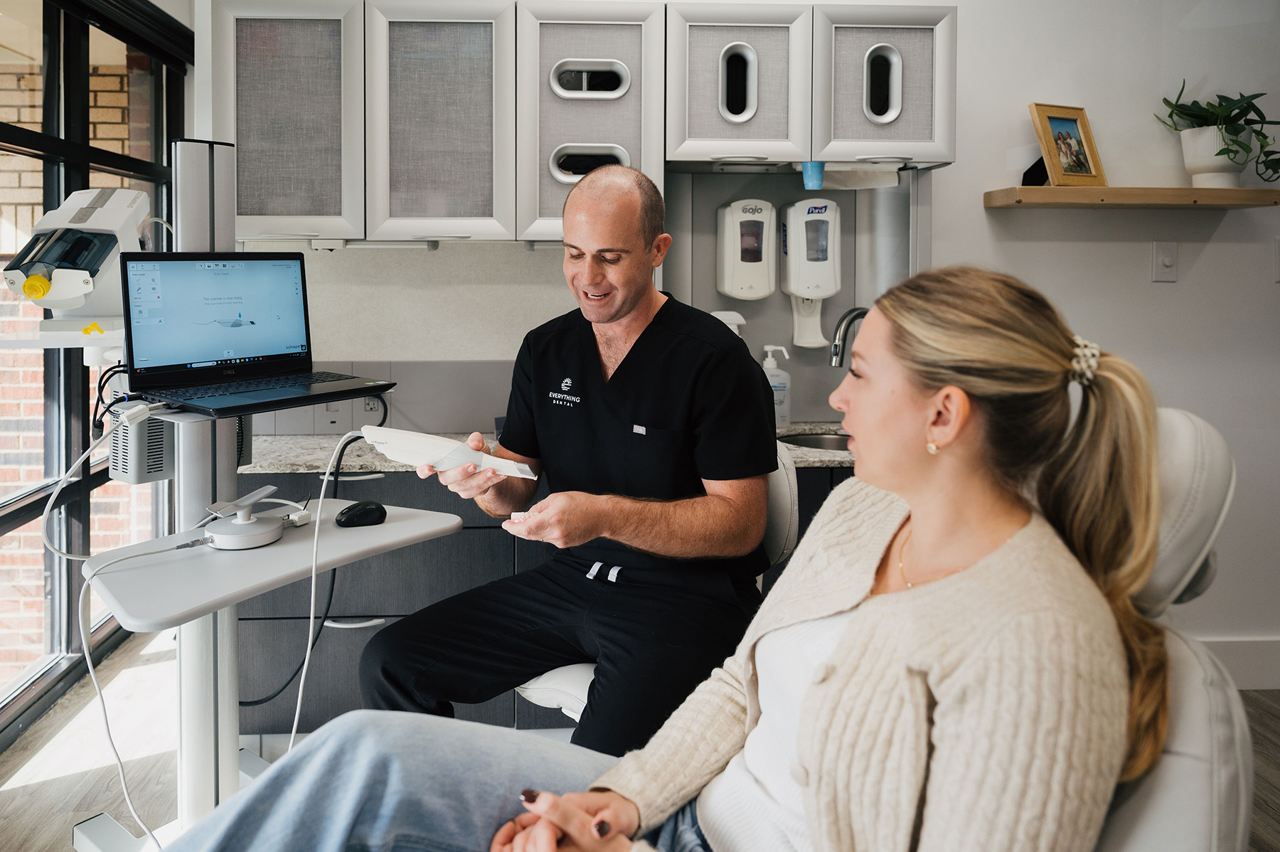 Dentist in black scrubs explaining a dental tool to a female patient seated in a dental chair.