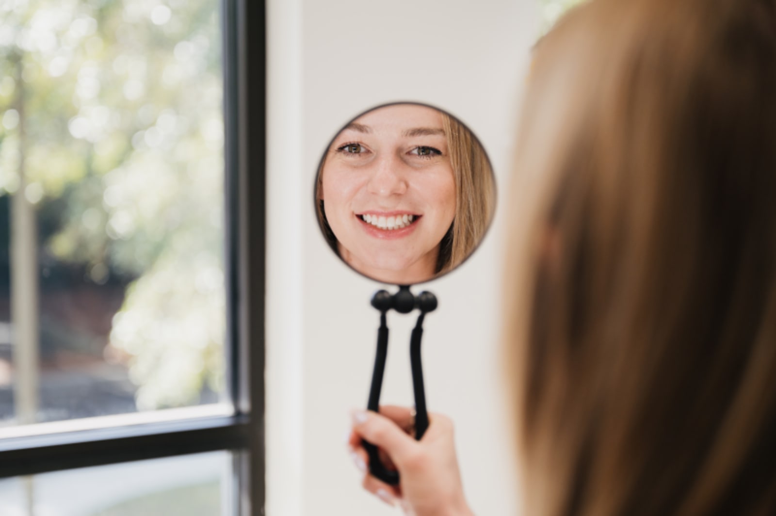 Woman holding mirror examining her smile indoors by a window.