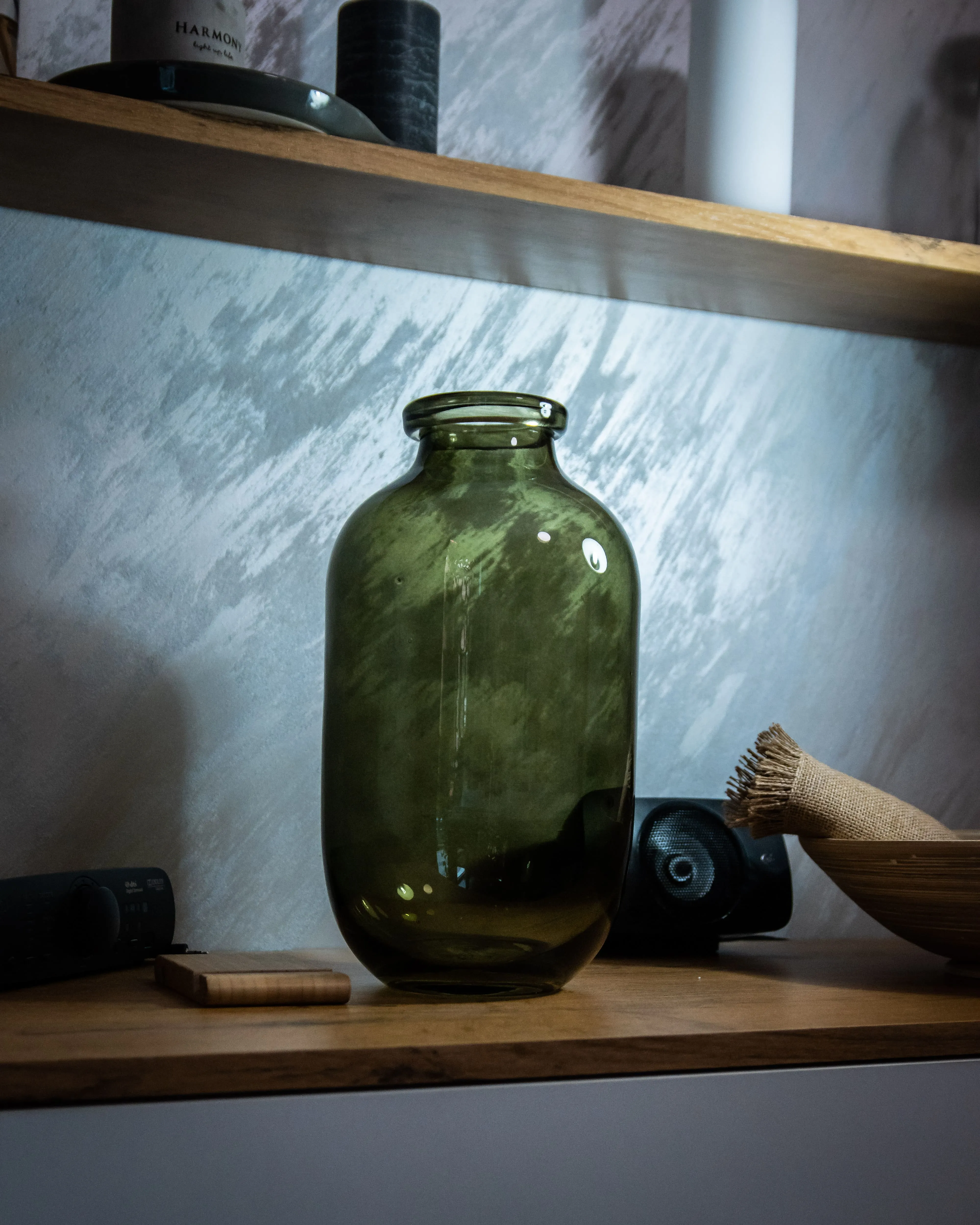 Dekorativna zelena staklena vaza na drvenoj polici kao deo modernog i prijatnog enterijera salona Runway Massage na Vračaru.