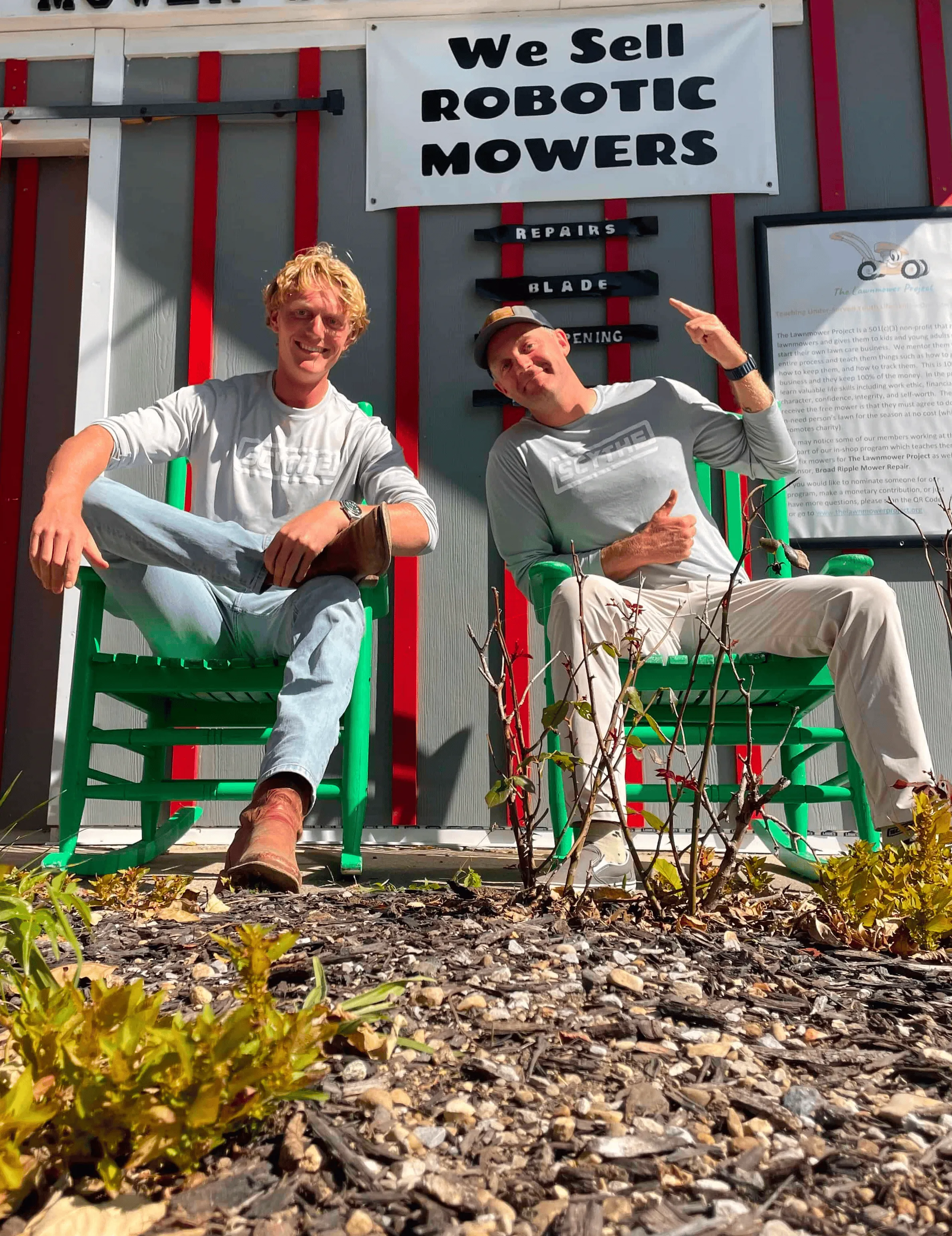 Two people sitting outside in green chairs. One is pointing to a sign behind him that says "We Sell Robotic Mower".