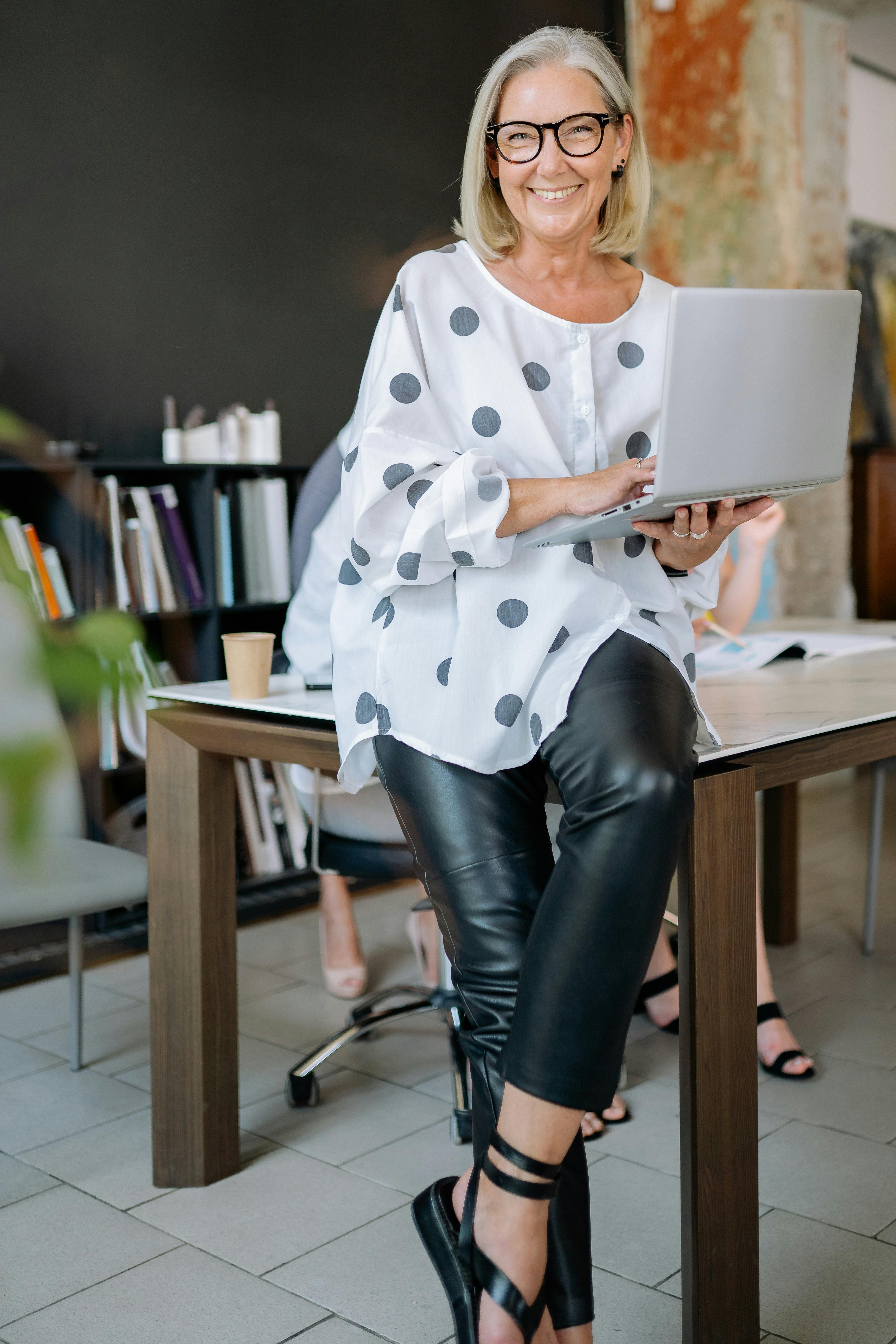 Stylish senior woman wearing polka-dot blouse and black leather pants smiles confidently while holding a laptop in a modern office setting.