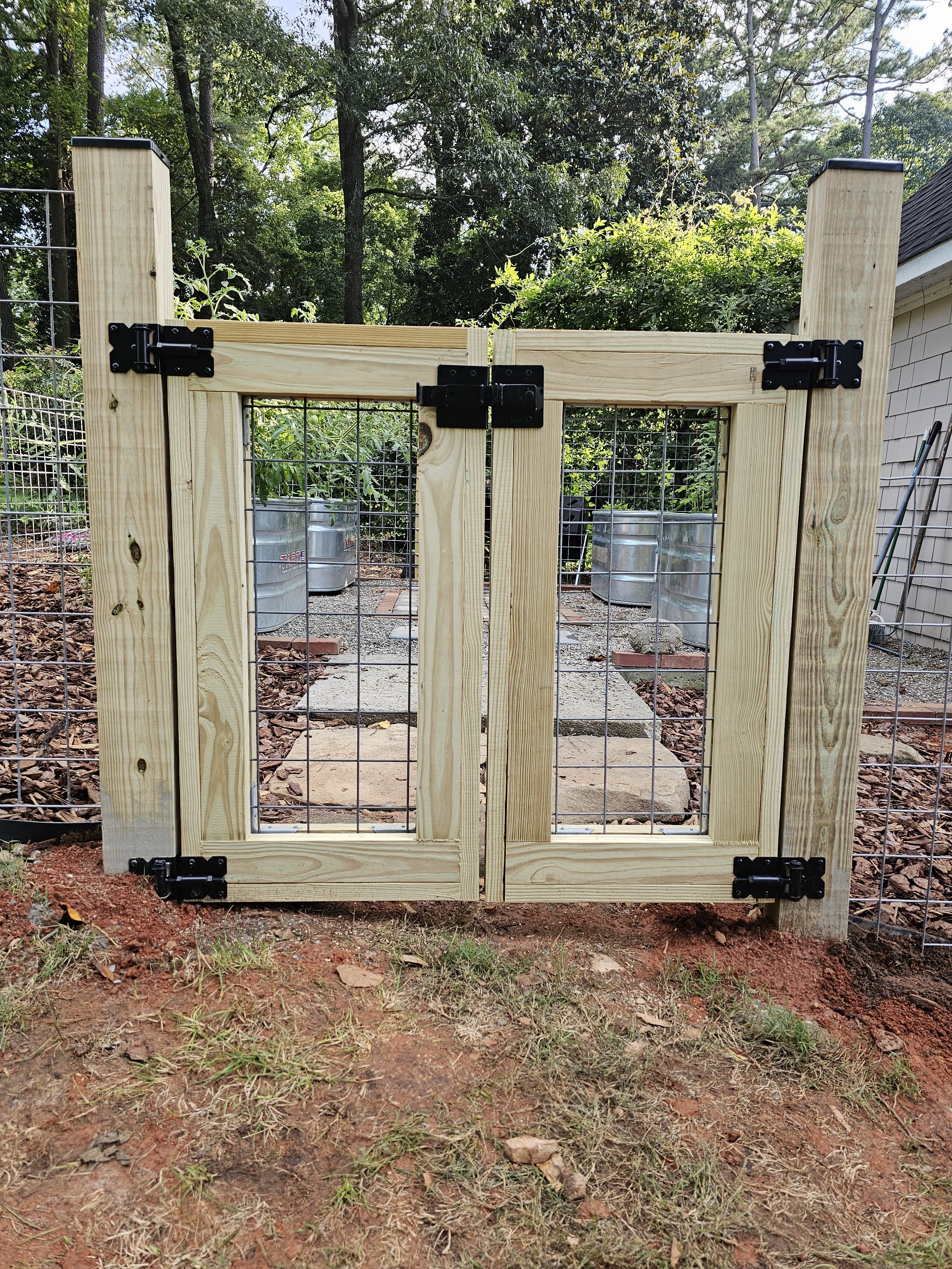 Wooden double garden gate with wire mesh panels and black metal hinges, set in a wire fence with a backyard garden visible behind.