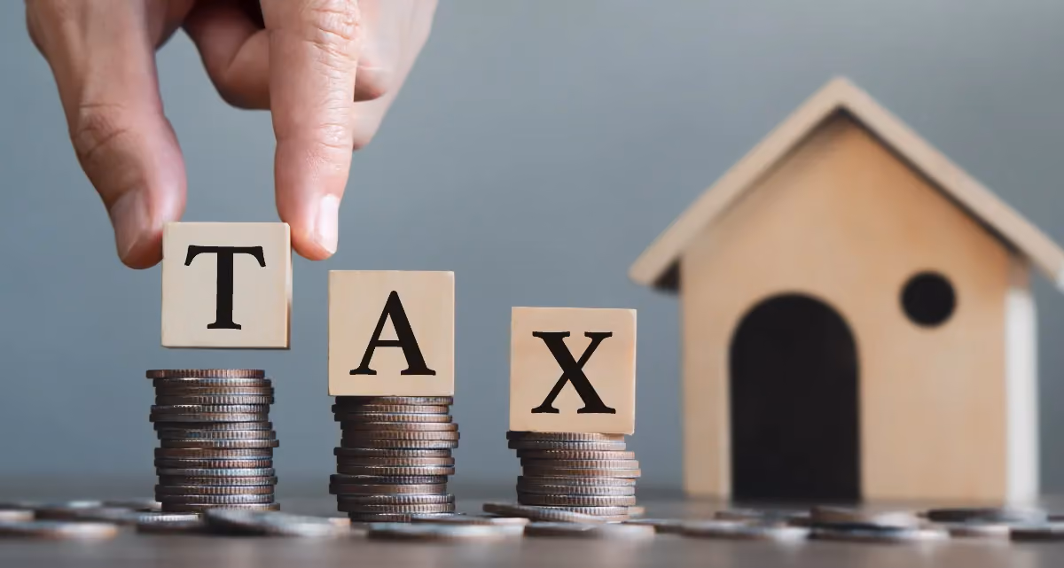 Three stacks of coins next to a model house with letter blocks marked T, A and X being placed on top of them