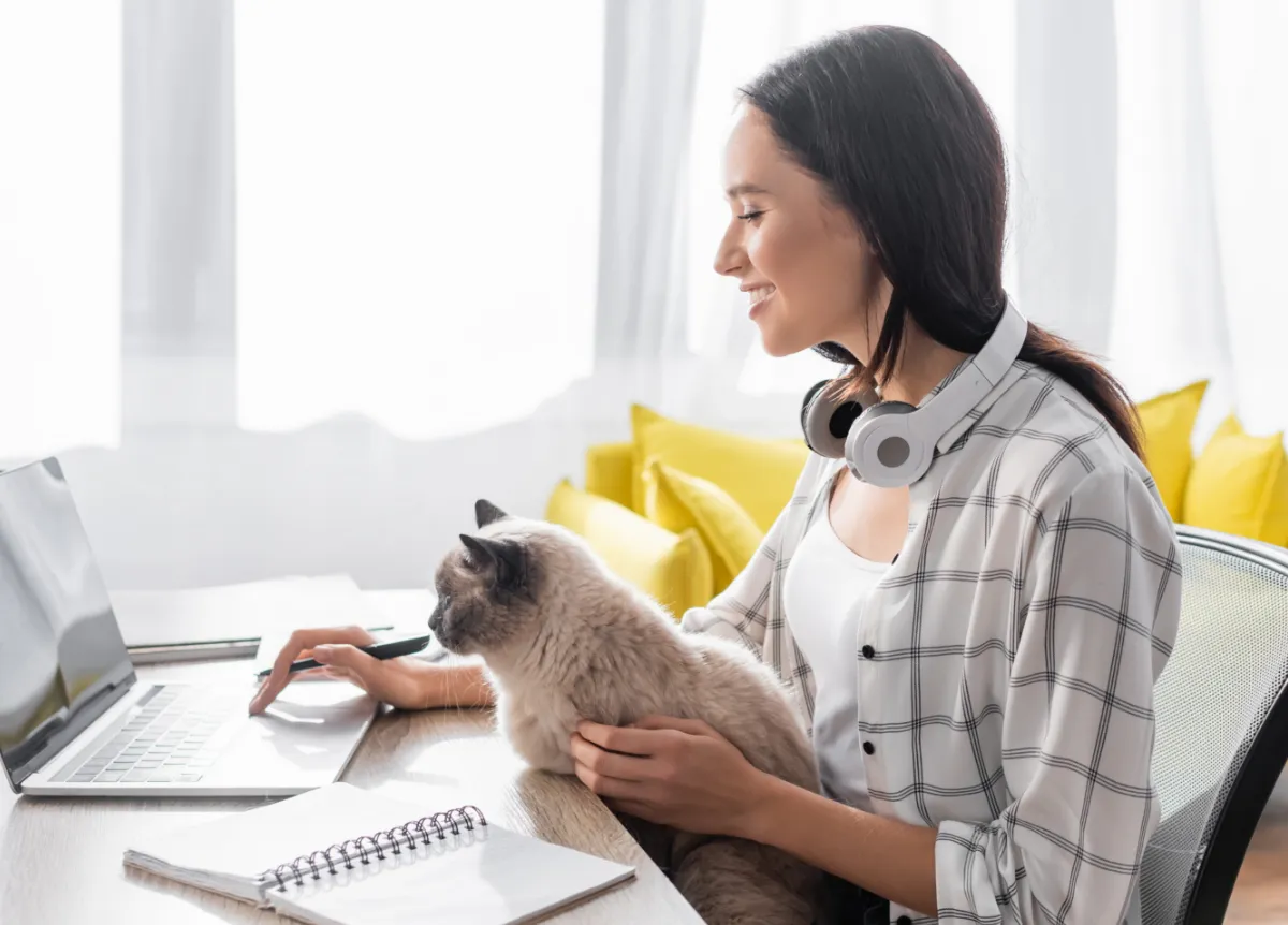 Smiling woman typing on laptop while sitting at desk with cat on her lap