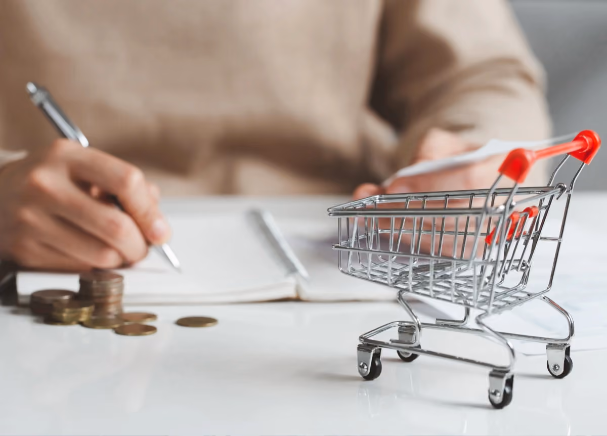 A woman makes her grocery budget with pen and paper with a shopping cart in the foreground.