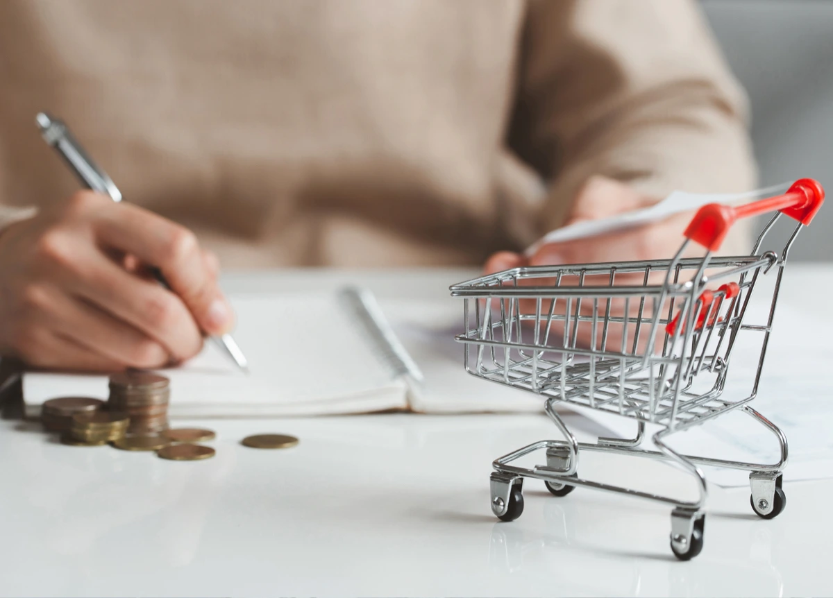 A woman makes her grocery budget with pen and paper with a shopping cart in the foreground.